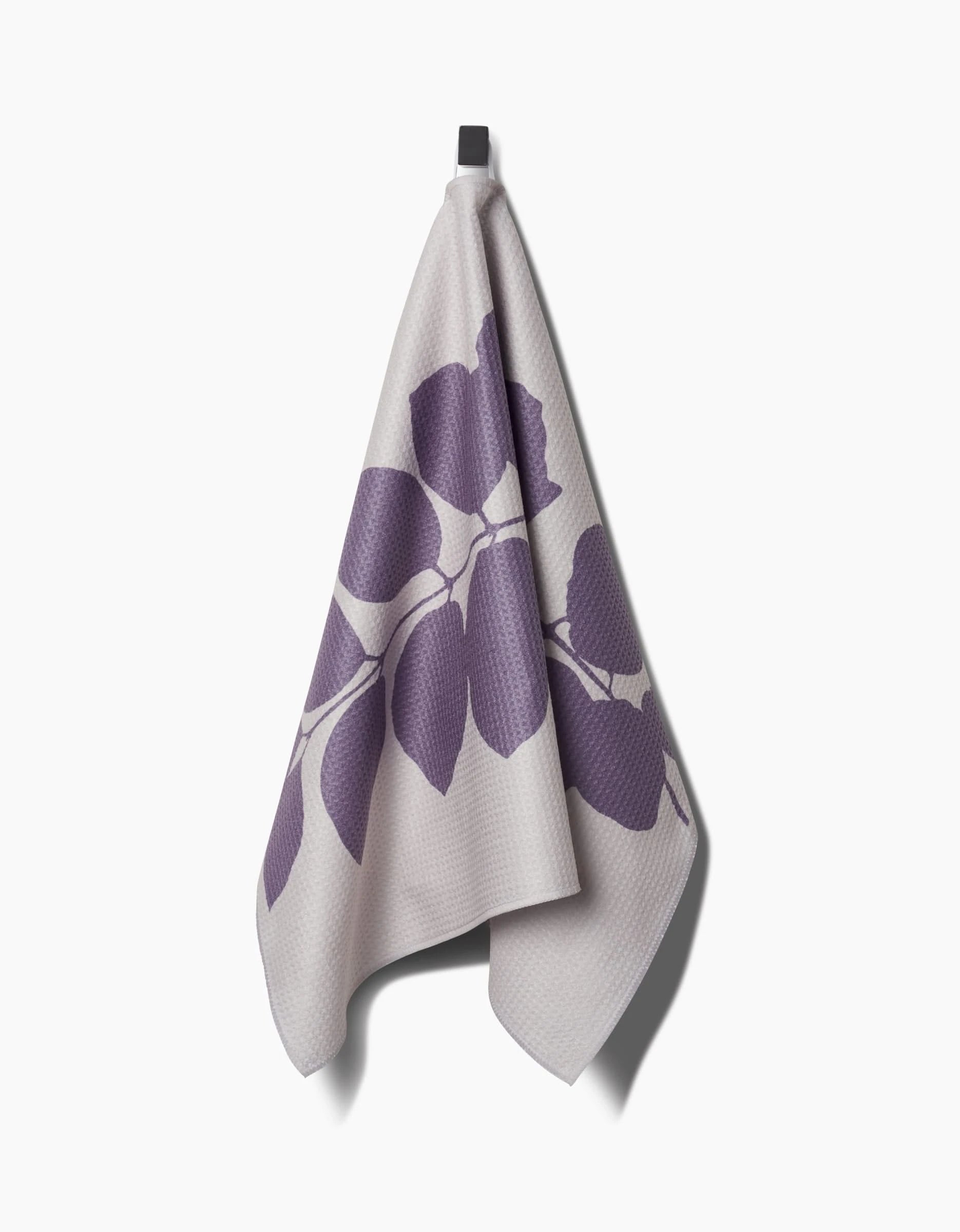 The Carob towel by PrintedPlant hangs on a hook, showcasing a bold purple leaf design on light gray. Made from ultra-absorbent Geoweave fabric, it offers both style and superior drying performance.