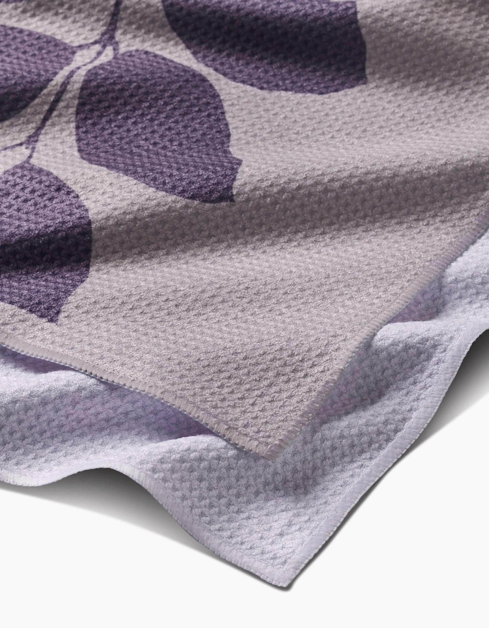 A close-up of the PrintedPlant Carob towel shows folded light purple Geoweave fabric with a dark purple leaf edge. Its dimpled, waffle-like texture is ultra-absorbent and adds a stylish touch.