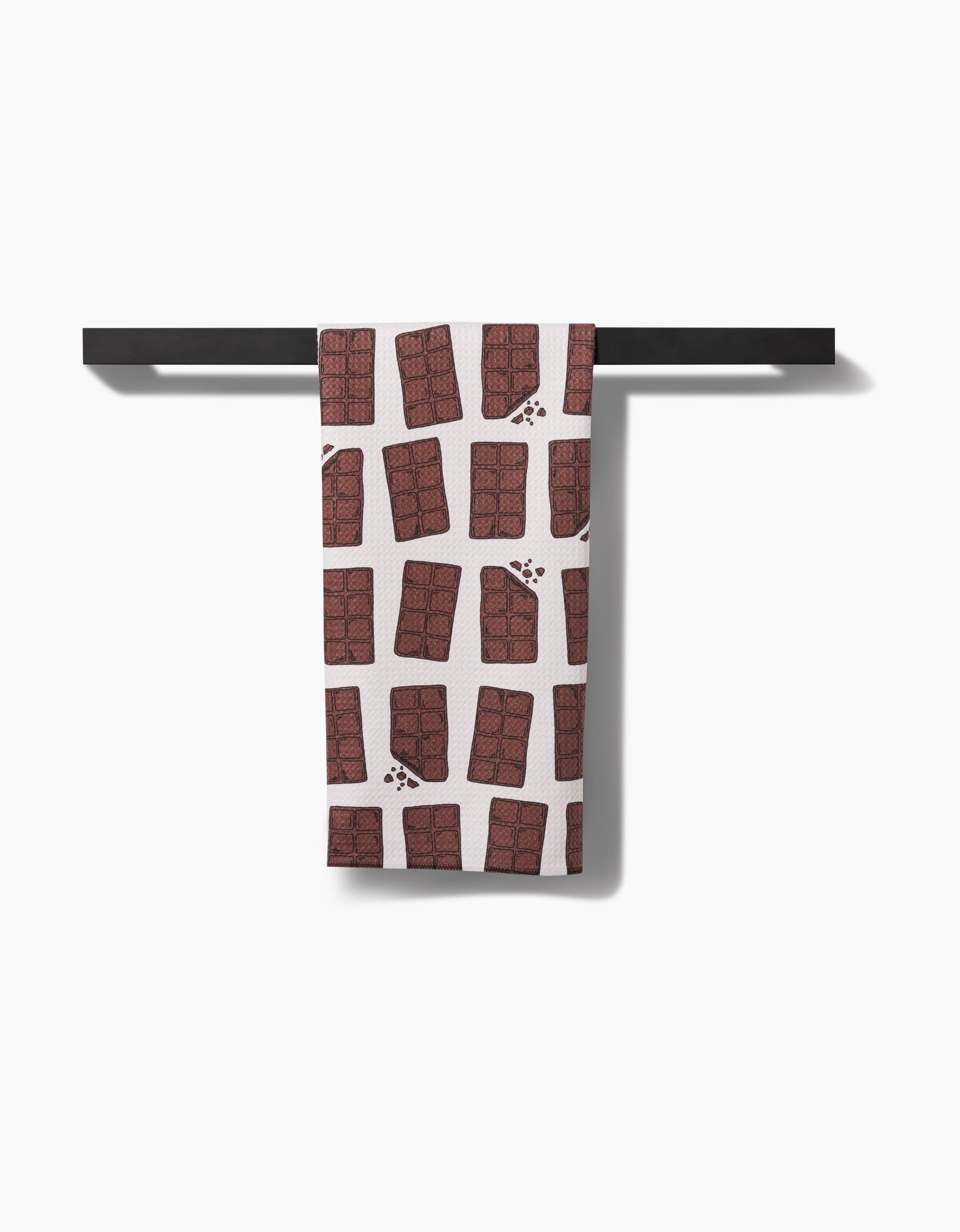 The Chocolate Everything tea towel by Amy Droubay, featuring brown chocolate bars on crisp white Geoweave fabric, hangs on a sleek black rack—combining style and sustainability.