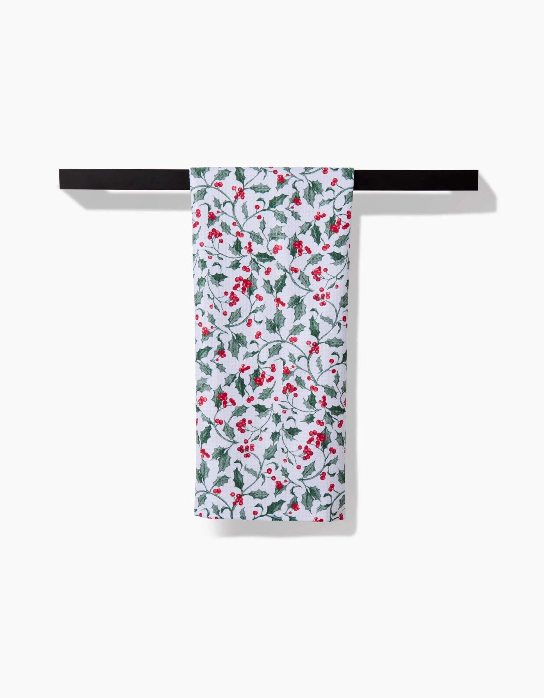 The Aubrey Fairchild Christmas Holly towel, featuring green holly leaves and red berries on white, hangs neatly on a black towel bar against a plain white background.