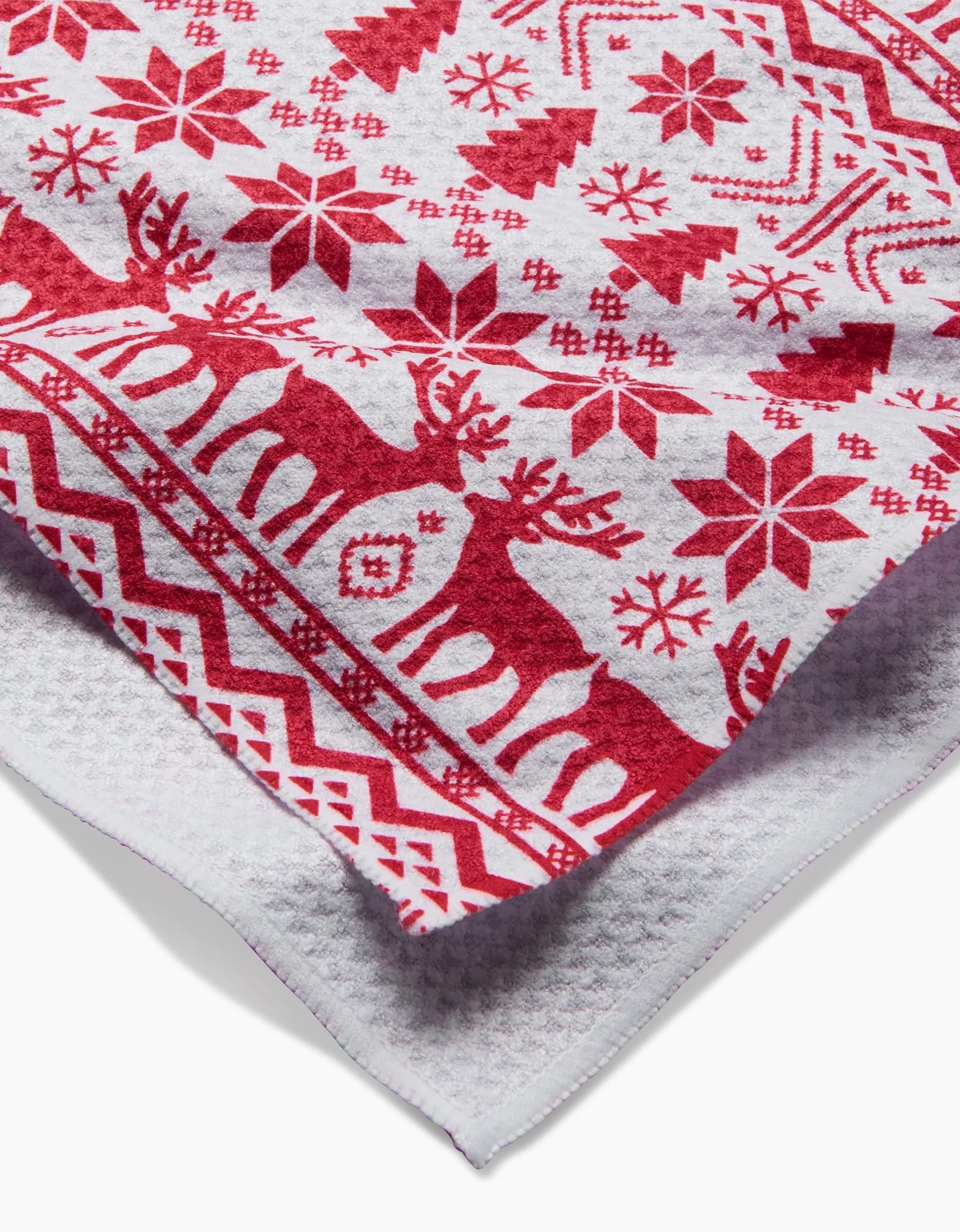 A close-up of the Hufton Studio Christmas Jumper tea towel in white, featuring red reindeer, snowflakes, stars, zigzags, and Christmas trees, with its fabric edge slightly folded.