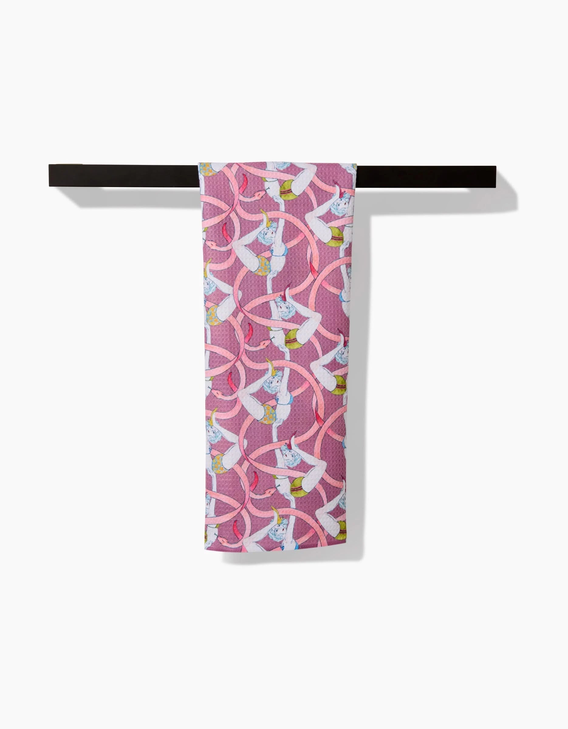 The Circus tea towel by Maayan Alper-Swan is pink, made from recycled materials, and displays a repeated pattern of white cats in sunglasses holding yellow drinks, shown draped over a black bar on a white background.