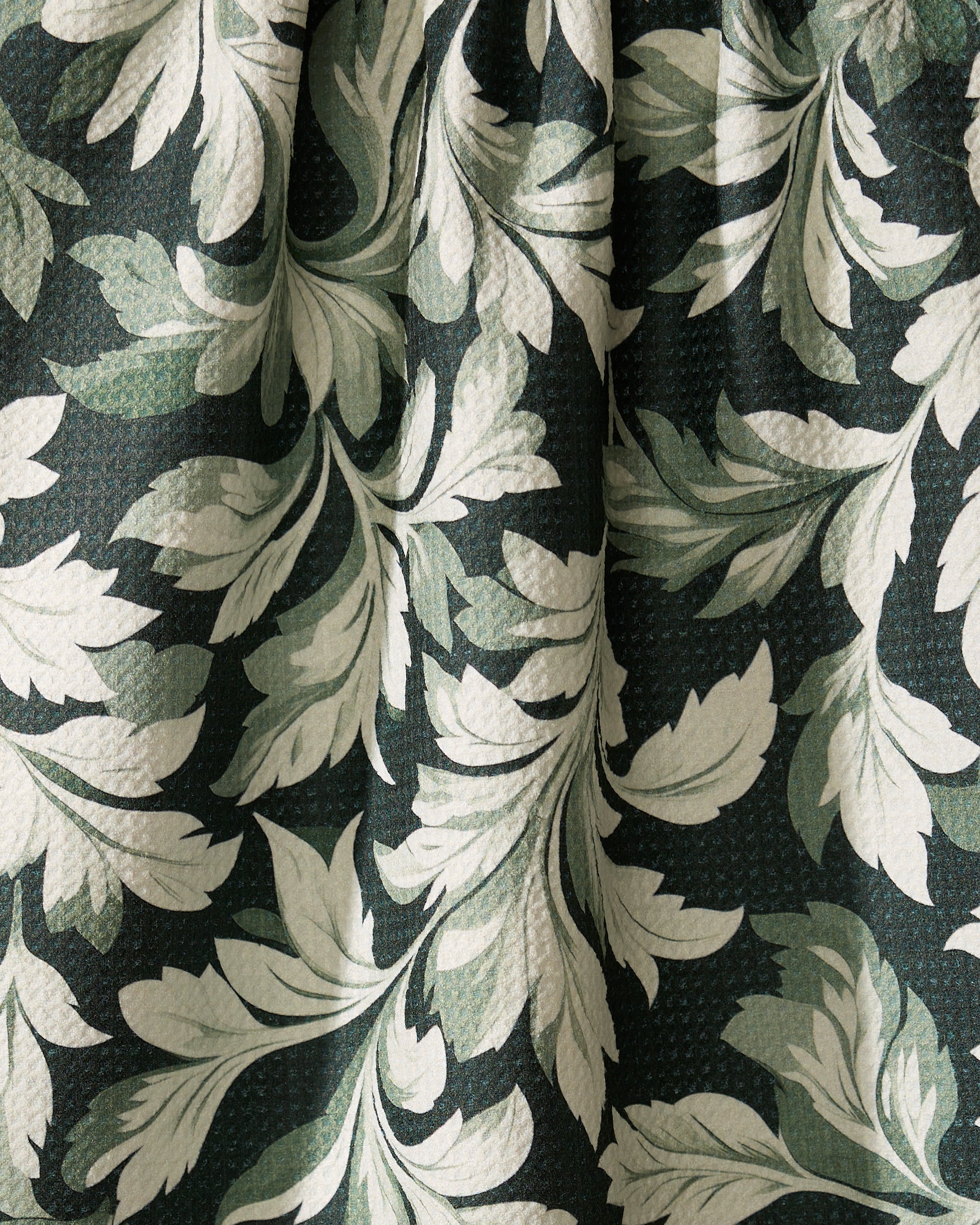 Close-up of Geometry’s Classic Foliage kitchen towel, showing dark green textured fabric with a botanical pattern of large cream and light green leaves. Made from recycled materials, its gentle folds enhance the leafy design.