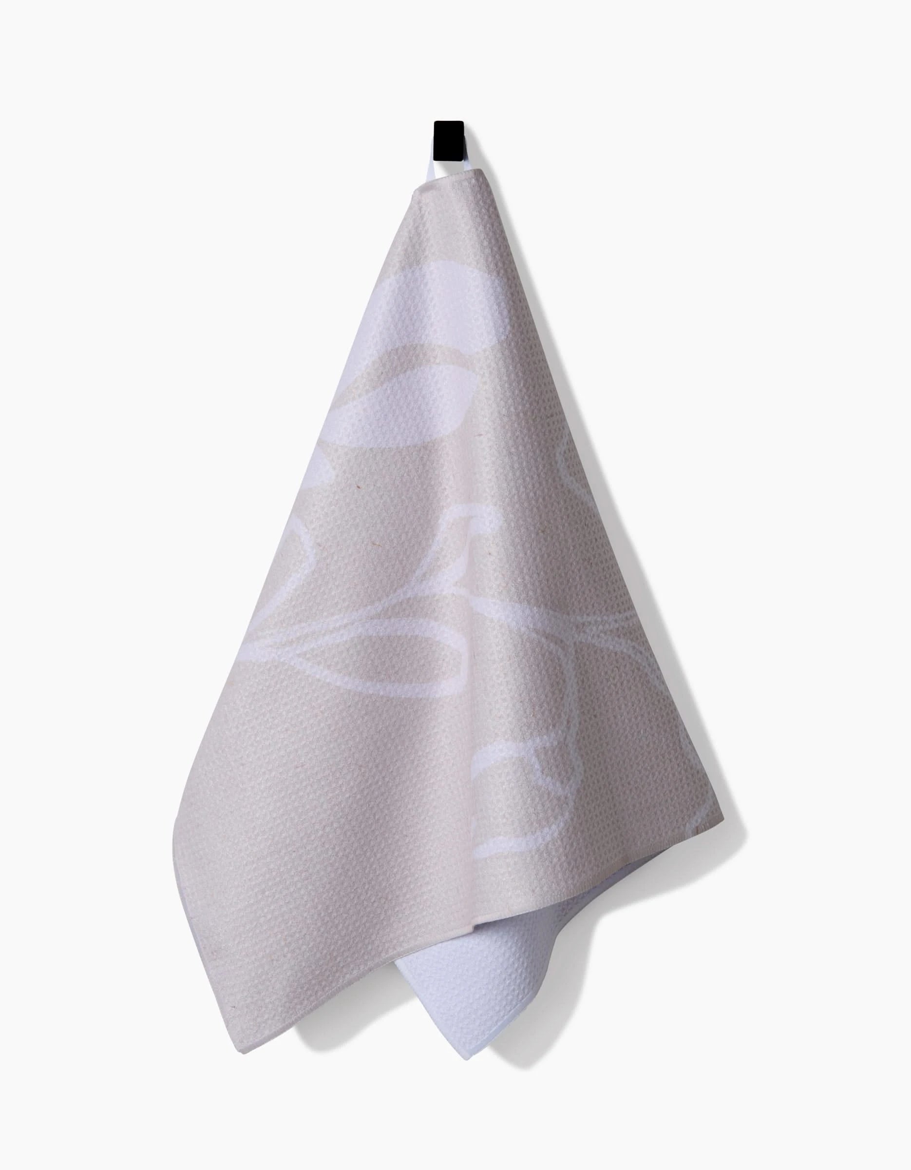 The GEOMETRY Clean tea towel, featuring an abstract beige and white design and made from recycled materials, hangs from a black hook against a plain white background.