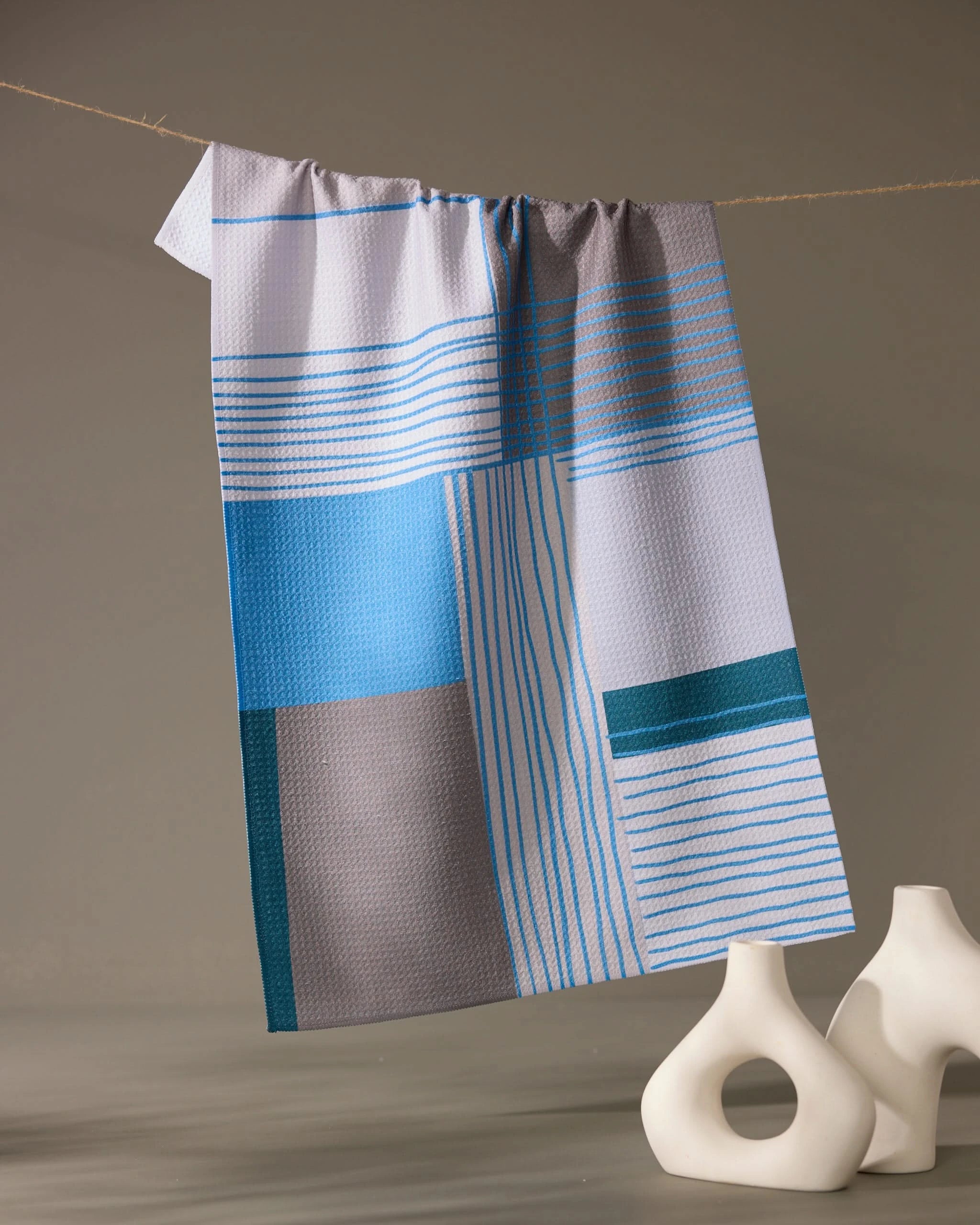 The Geometry “Cool Blocks” recycled kitchen towel, featuring a blue, gray, and white pattern, hangs on a string against a neutral background with two modern abstract white vases positioned to its right.
