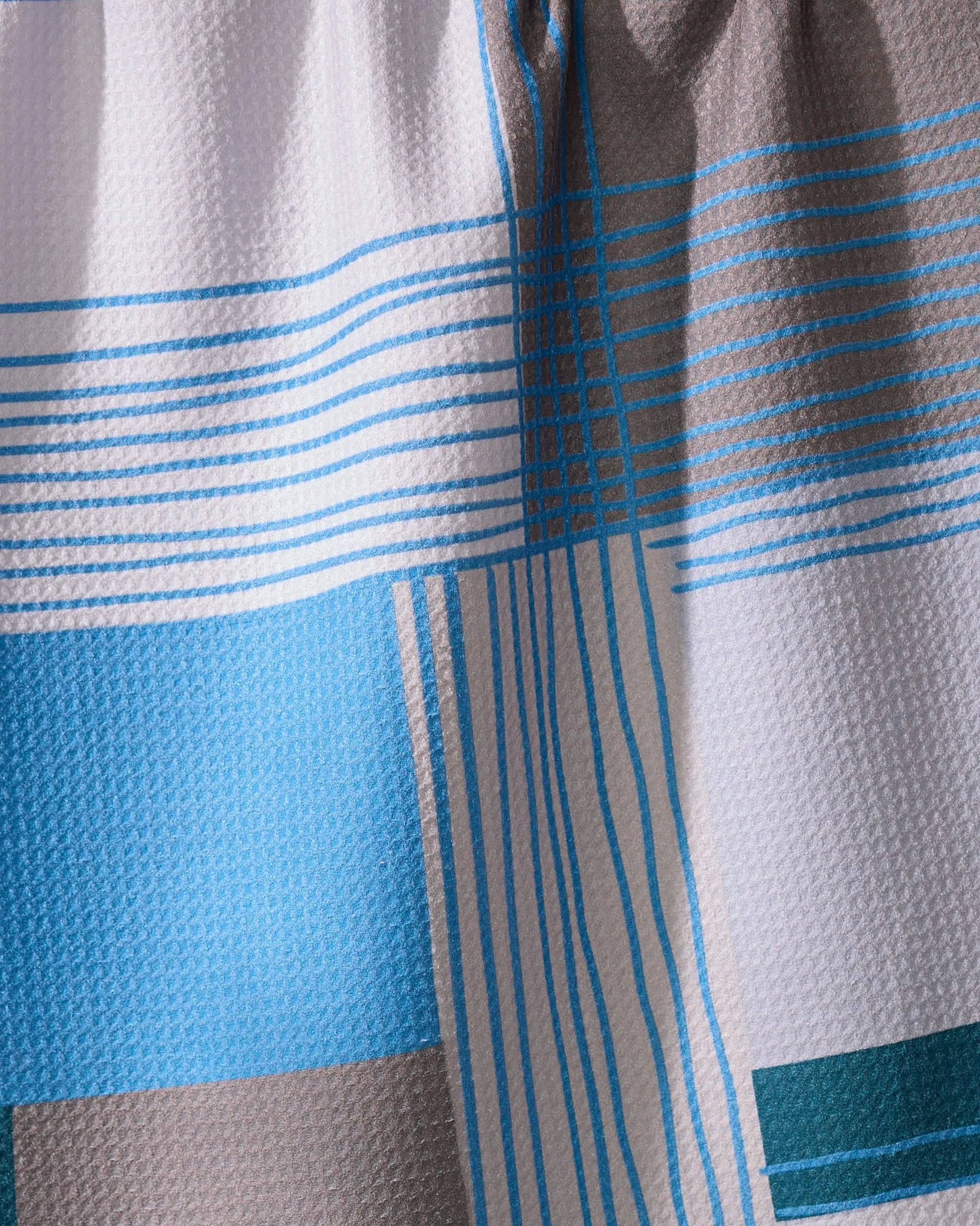 A close-up of Geometrys Cool Blocks towel, showcasing a quick-drying fabric with a geometric blue, gray, and white block pattern accented by intersecting blue lines. The material is slightly gathered at the top.