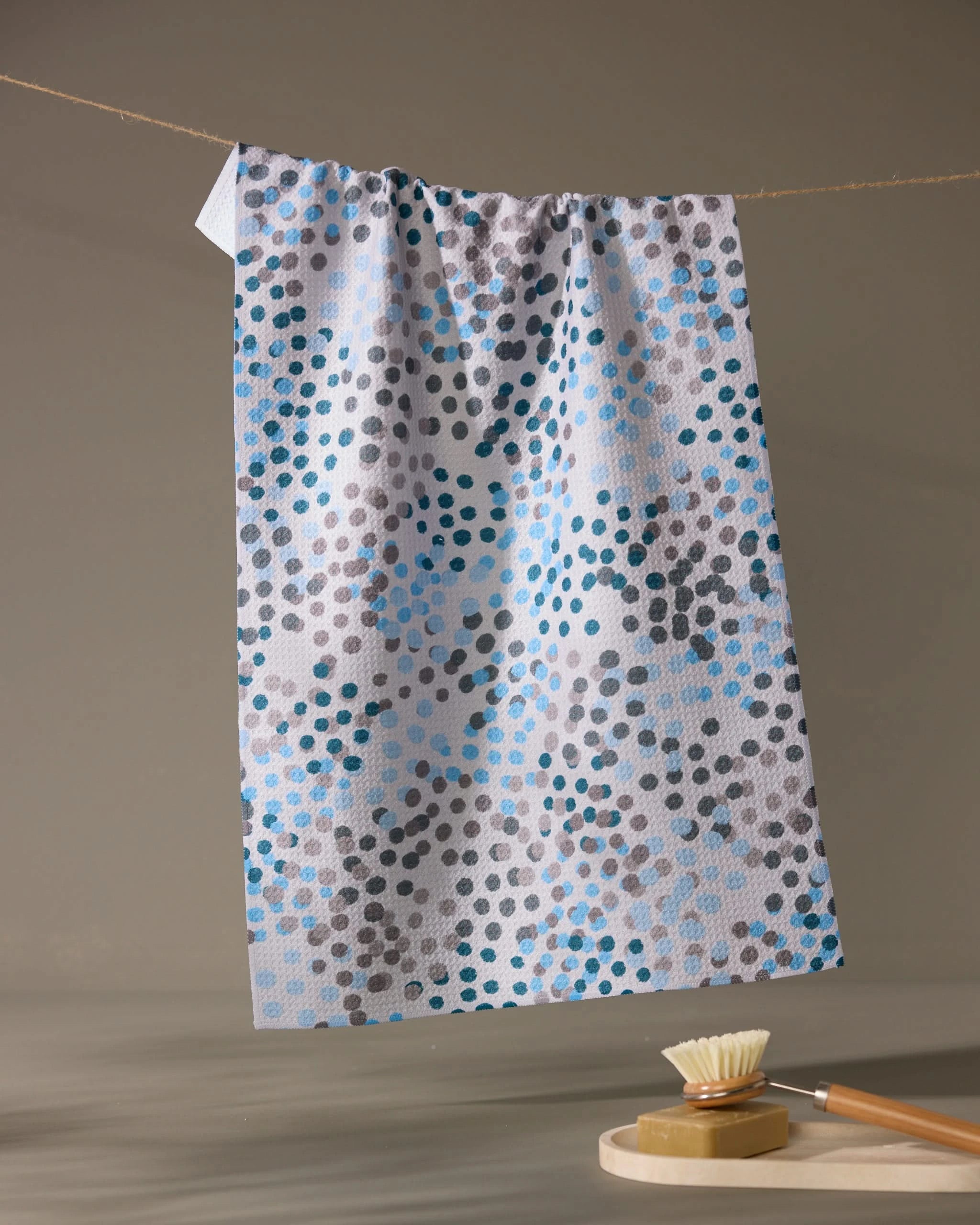 The Geometry Cool Dots ultra-absorbent white kitchen towel with blue, gray, and black dots hangs on a clothesline above a wooden brush with light bristles resting on a round soap dish in a neutral, softly lit setting.