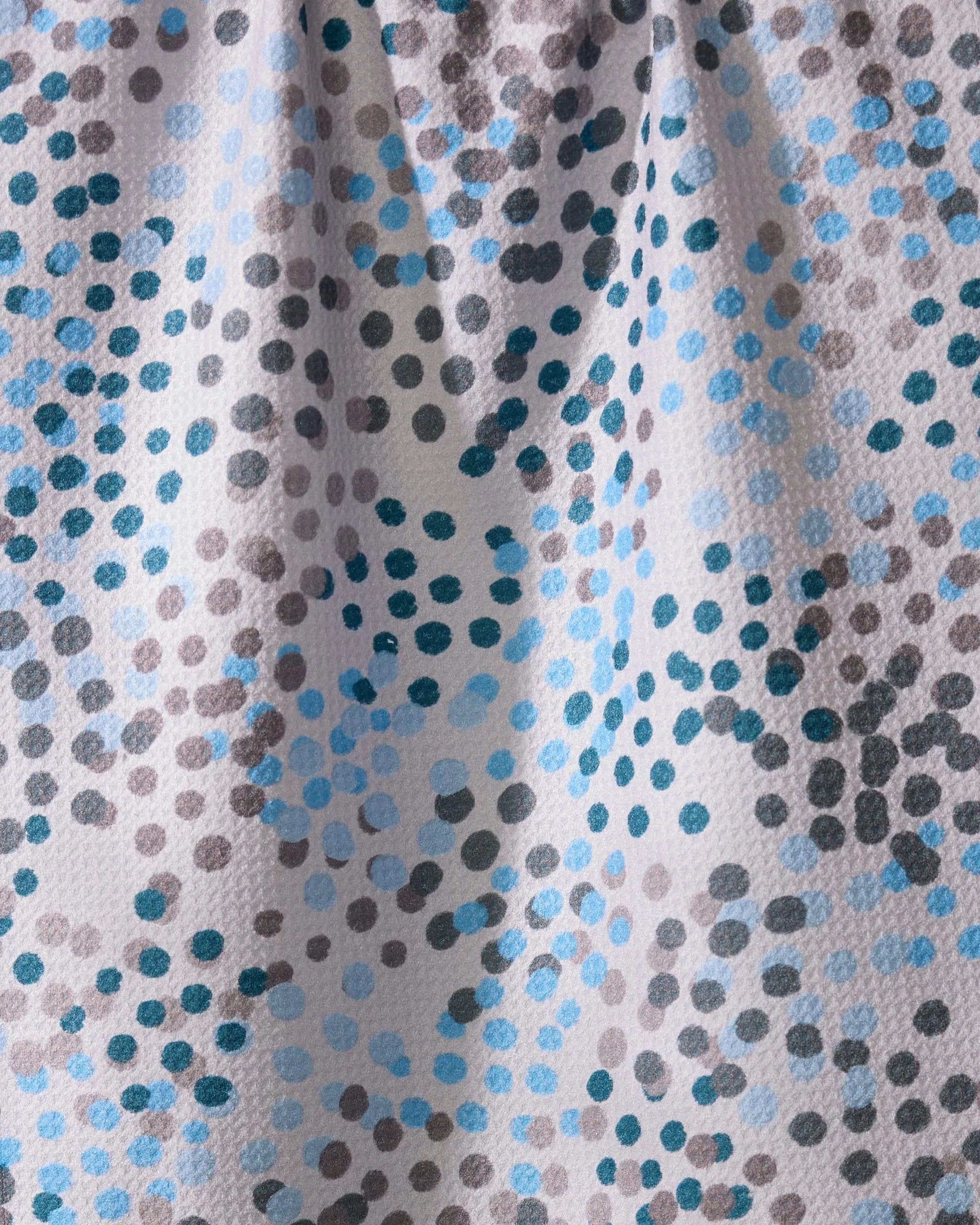 A close-up of Geometry’s Cool Dots kitchen towel shows its textured, ultra-absorbent recycled fabric with an abstract pattern of blue, gray, and black dots scattered on white, highlighted by gentle folds and shadows.