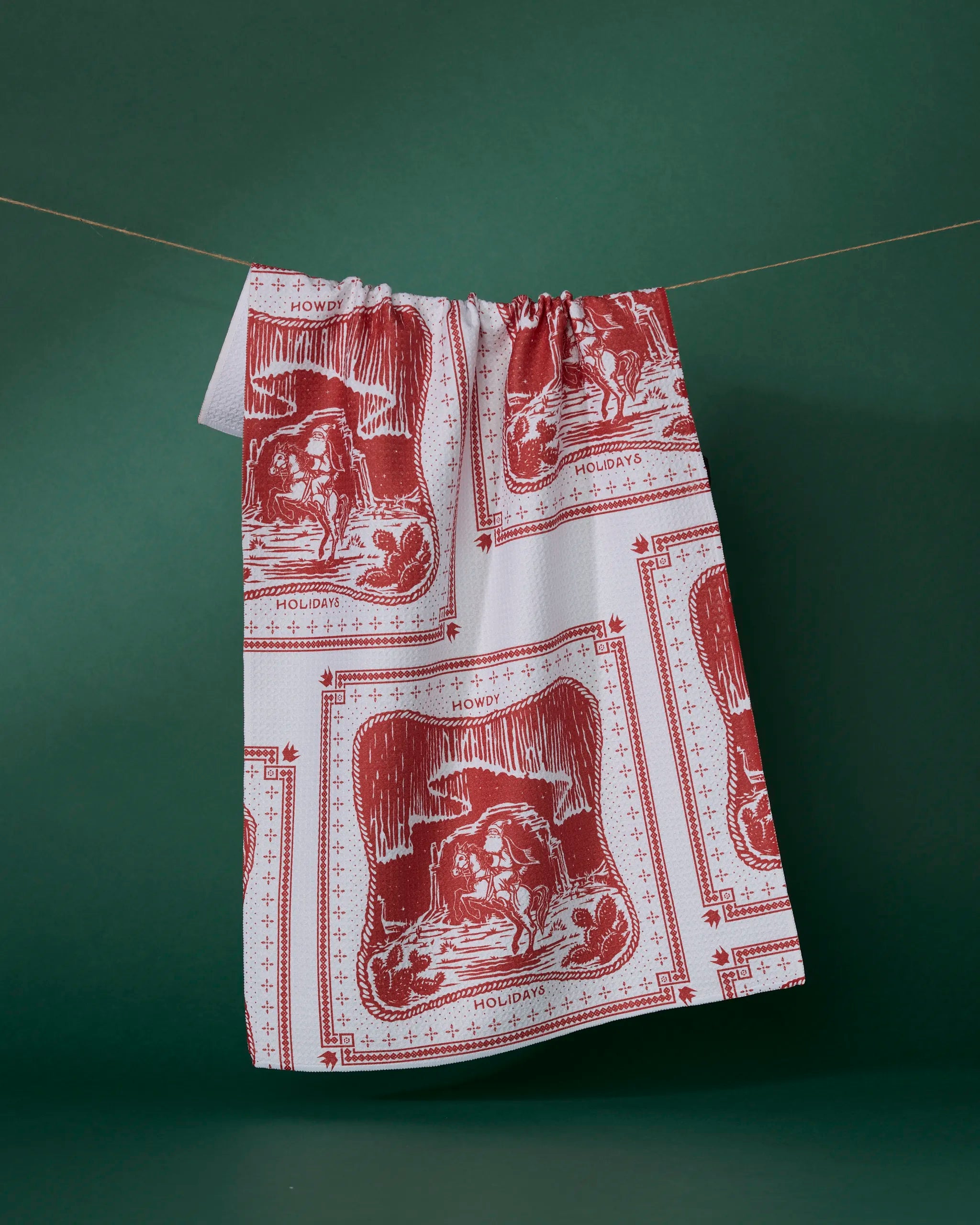 A white and red "Cowboy Christmas" tea towel by Geometry hangs on a line against a dark green background, made from recycled materials and featuring western-themed designs with a cowboy hat, boots, and "Howdy" above "Happy Holidays.