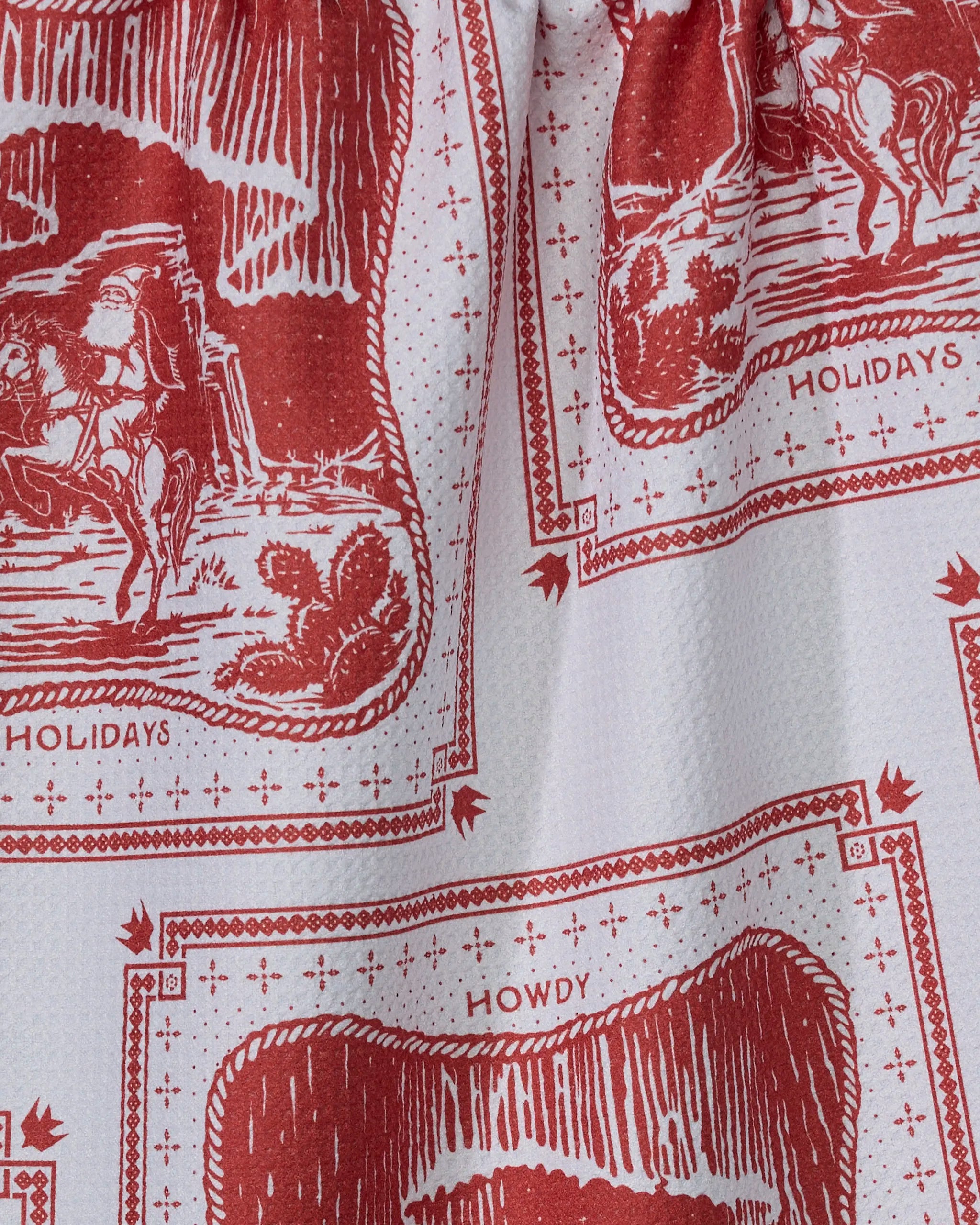 The Geometry Cowboy Christmas tea towel is ultra-absorbent, made from recycled materials, and features red and white Western prints with cowboys, “HOLIDAYS,” “HOWDY,” and decorative borders.