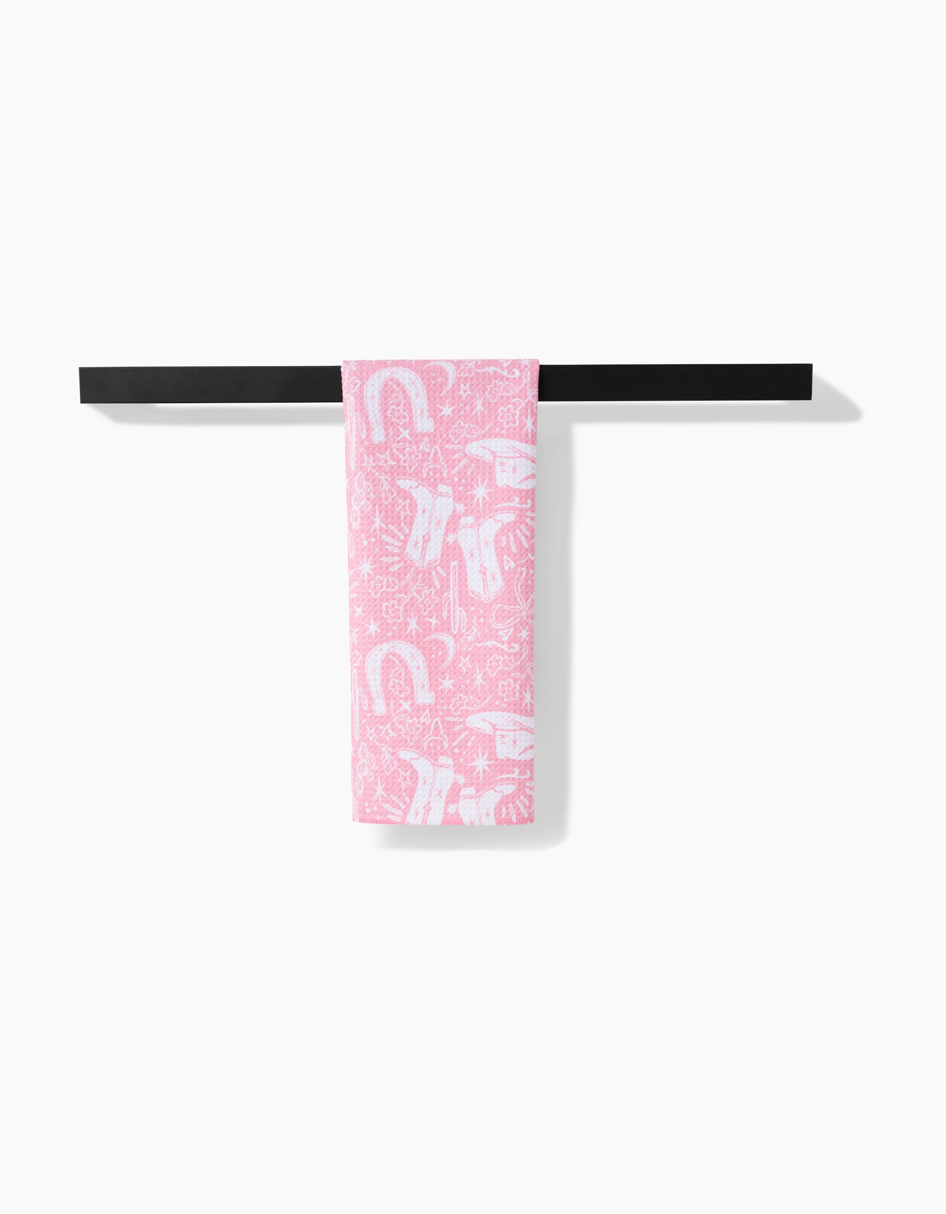 The Cowgirl Chic towel, pink with white boots, horseshoes, and stars, hangs neatly folded over a black rack. Made from ultra-absorbent Geoweave fabric, it stands out against a white background.