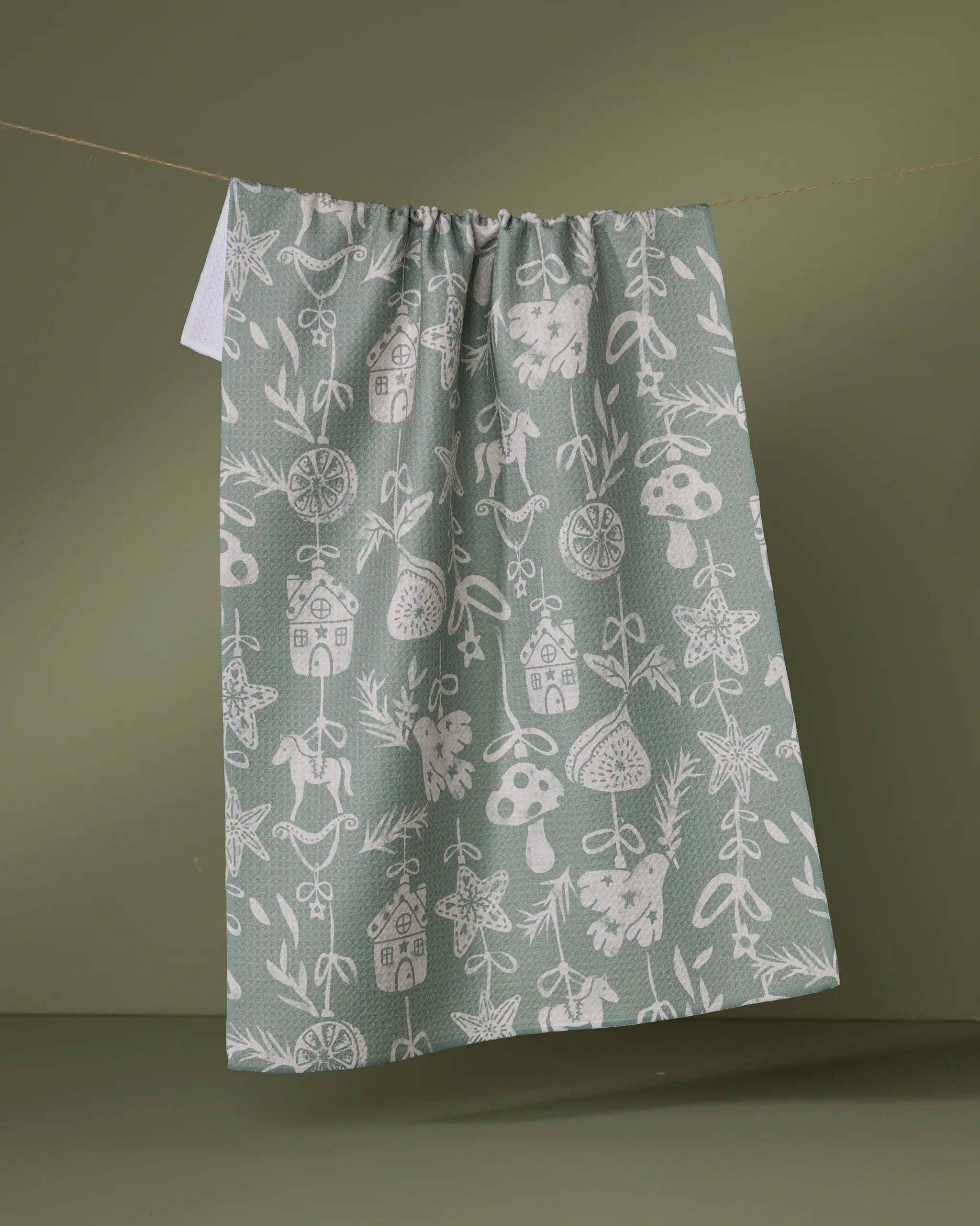 The Geometry "Cozy Ornaments" kitchen tea towel features white whimsical illustrations of mushrooms, houses, leaves, bows, stars, and citrus on green fabric made from recycled materials. Shown hanging against an olive background.
