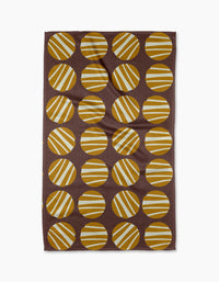 The Crazy Circles tea towel by Lizzie Clark is brown and rectangular, featuring a grid of evenly spaced yellow and white striped circles with diagonal stripes on its ultra-absorbent surface.