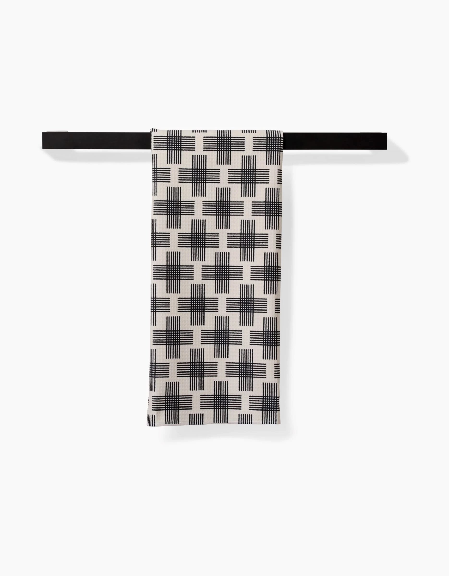 The Rosa Kichline Cross Hatch towel rack in black holds a white tea towel with a black geometric crosshatch square pattern, crafted from recycled materials, against a plain white background.