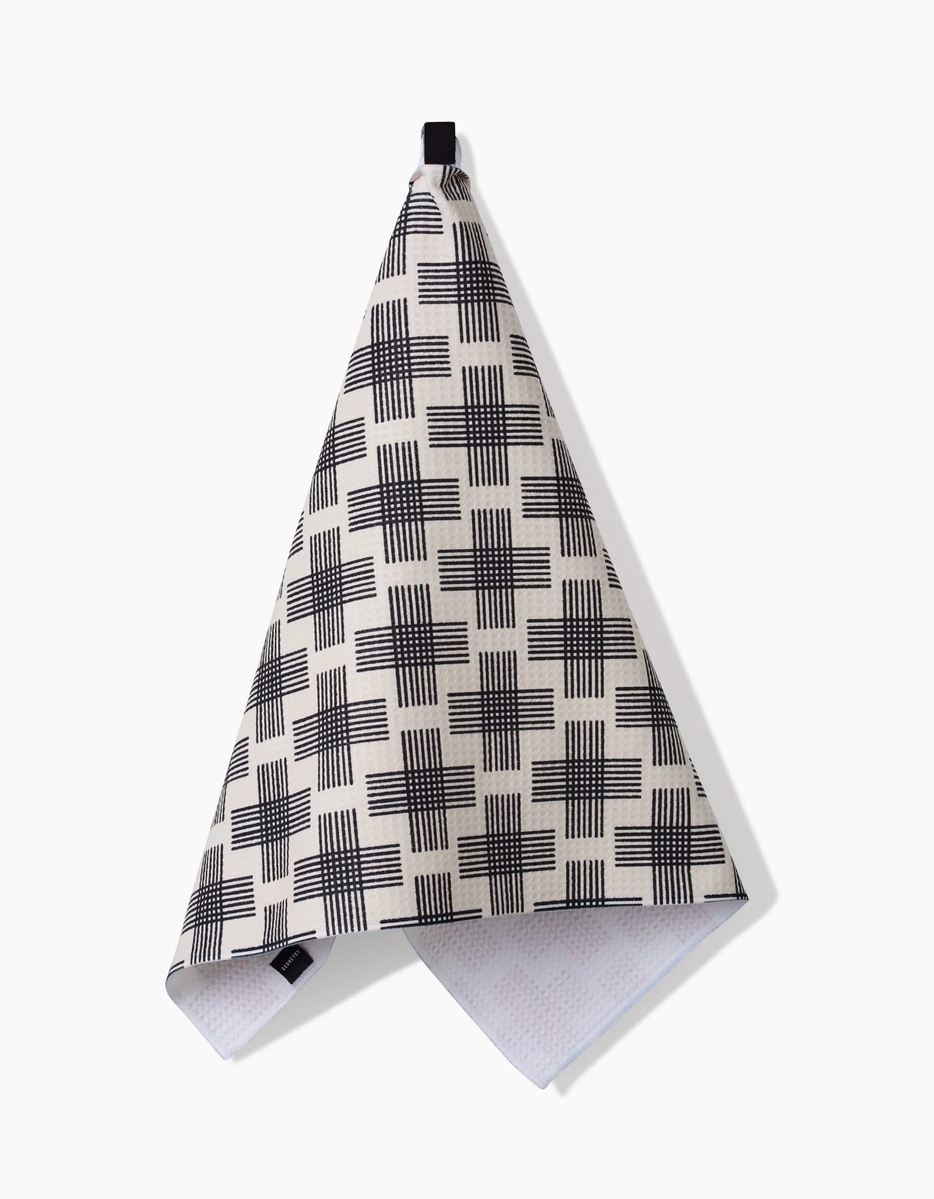 The Rosa Kichline Cross Hatch tea towel, made from beige Geoweave fabric with a black crisscross grid pattern, hangs by a black loop against a white background.