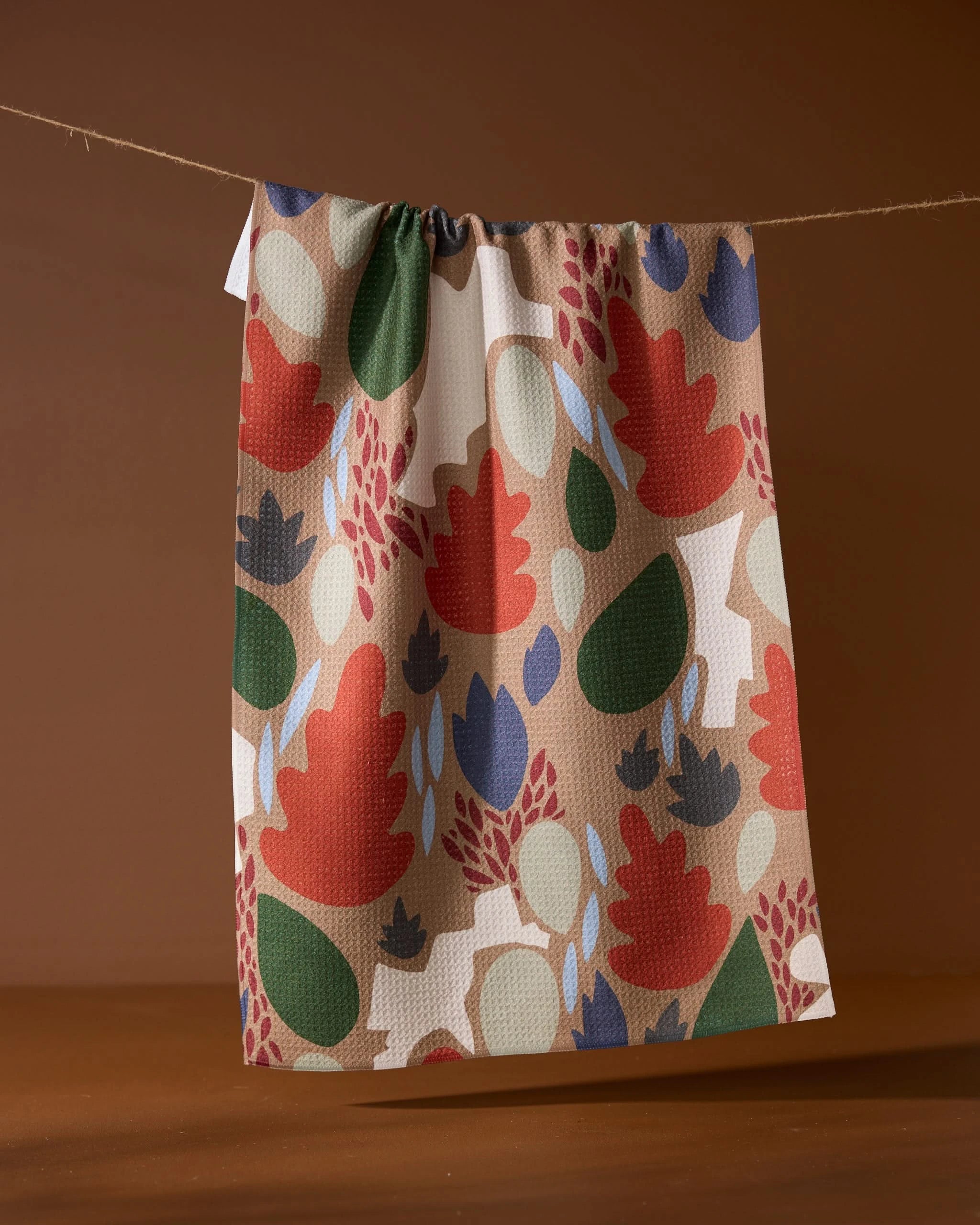 The Geometry Cut Paper Leaves Mocha tea towel, featuring abstract red, green, navy, and beige foliage shapes, hangs on a string against a plain brown background.