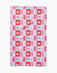 The Daily Icons tea towel by Laura Supnik is rectangular, made from recycled materials, and features a grid of colorful hearts, rainbows, flowers, suns, and abstract shapes in red, pink, blue, and white.