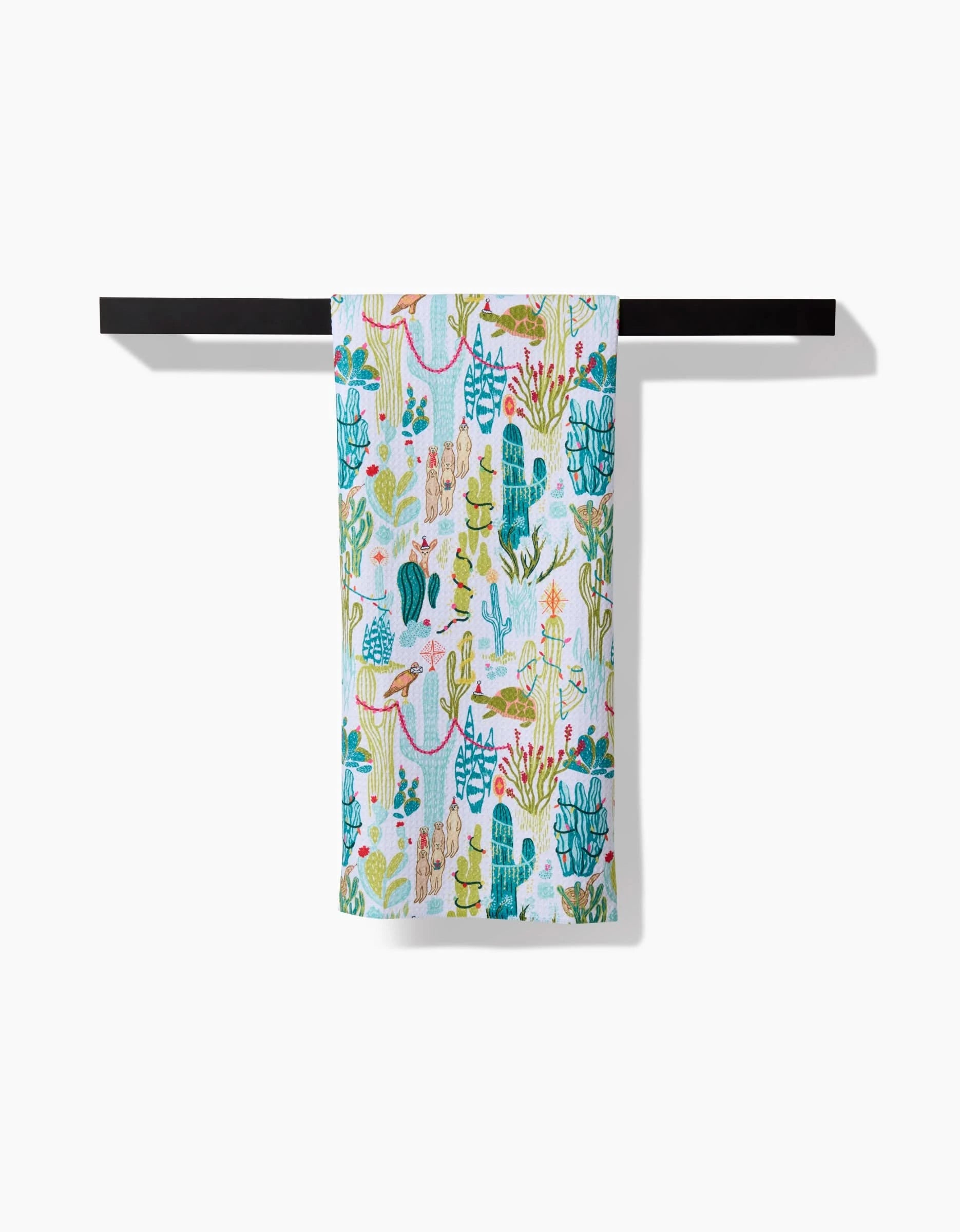 The Desert On Holiday kitchen towel by Elisabeth Wing, featuring vibrant cacti, plants, and llamas in green, blue, and pink, hangs neatly on a black towel bar against a white background.