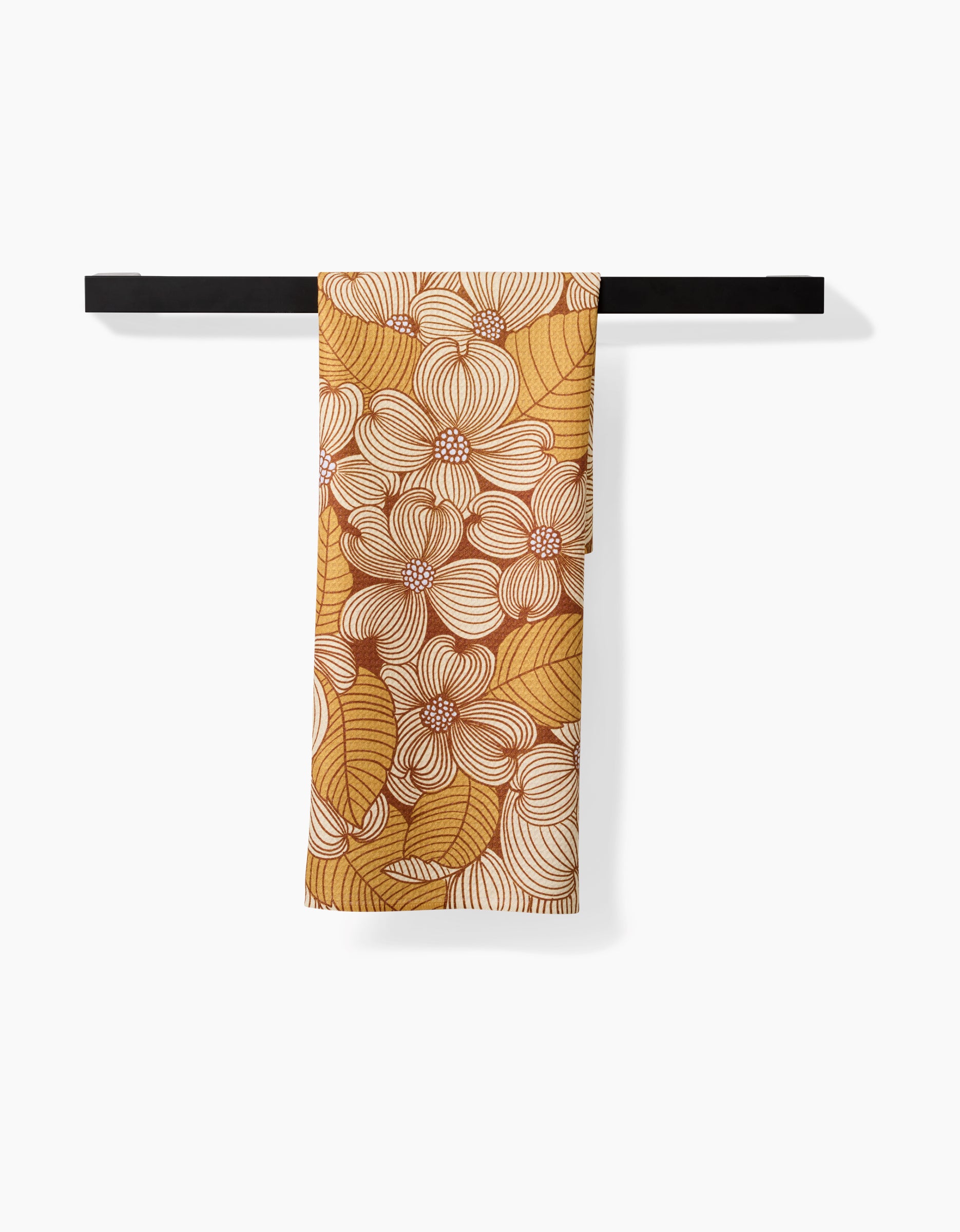 The Dogwood tea towel by Katie OShea, featuring a white and brown floral and leaf pattern on yellow and made from recycled materials, hangs folded over a black towel bar against a white background.