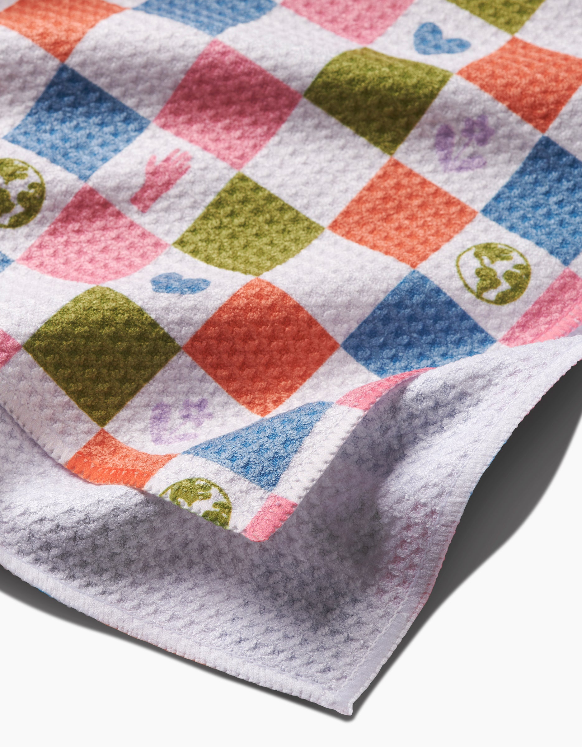 A close-up of Kayla Anns Earth Day Checkers tea towel, ultra-absorbent and made from recycled materials, with a textured white surface and a colorful checkered pattern of hearts, peace signs, and Earth symbols.