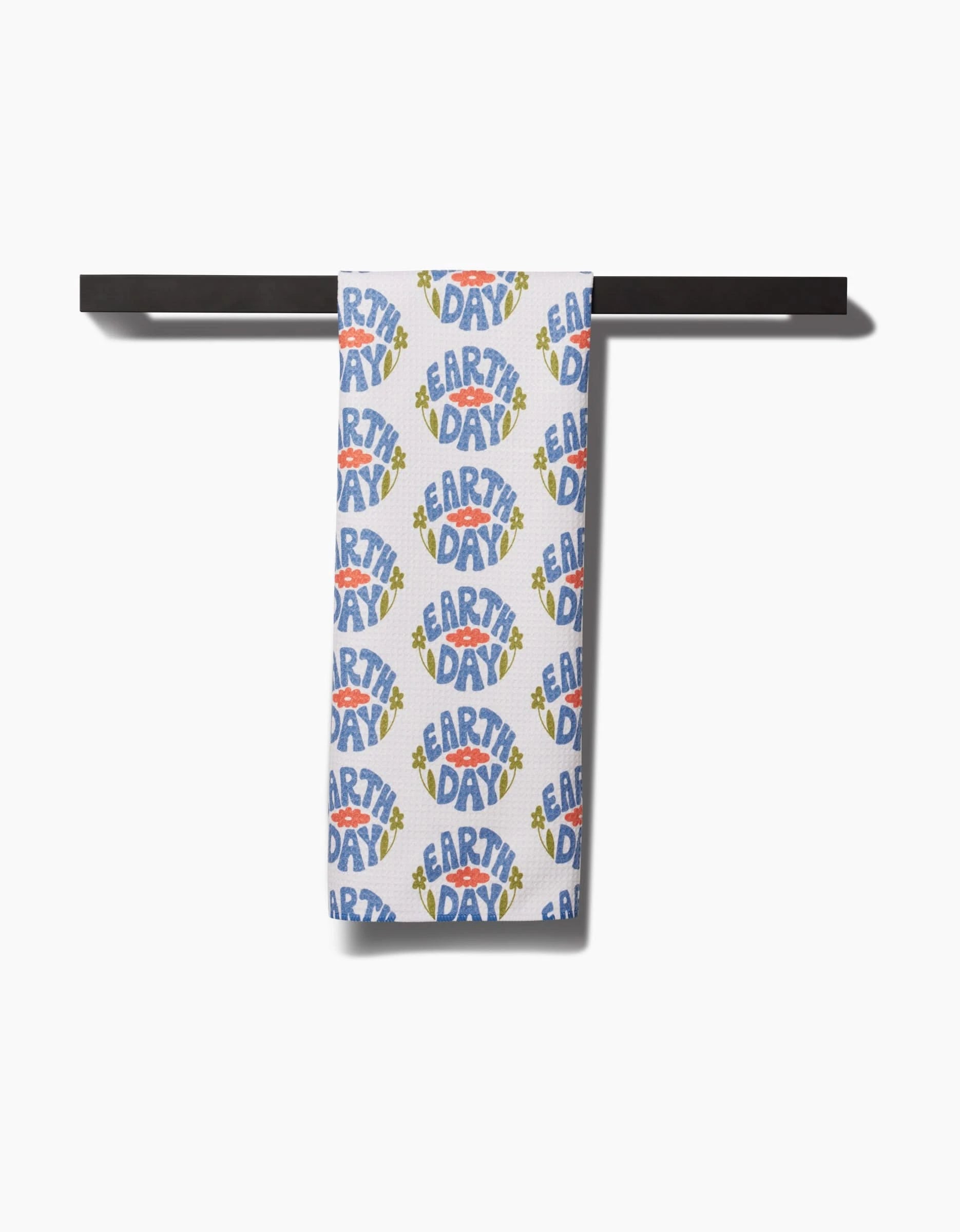 A white Kayla Ann Earth Day Stamp kitchen towel hangs on a black bar, featuring blue, yellow, and orange EARTH DAY text with green leaves and earth icons. Made from recycled materials.