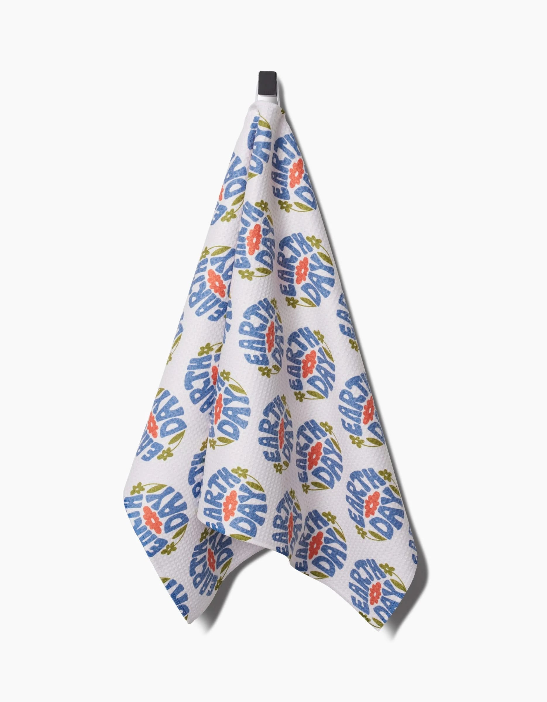 The Kayla Ann Earth Day Stamp tea towel hangs from a black hook, featuring blue and orange Earth Day-themed text and floral designs. Made from recycled materials, it brings an eco-friendly accent to your kitchen.