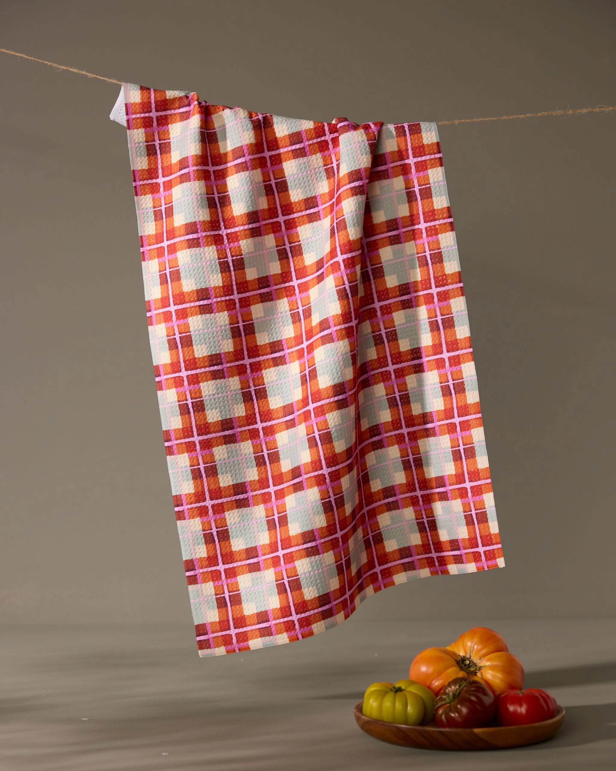 The Geometry Ecstatic For Autumn tea towel hangs from a clothesline above a plate of colorful tomatoes, set against a neutral background.