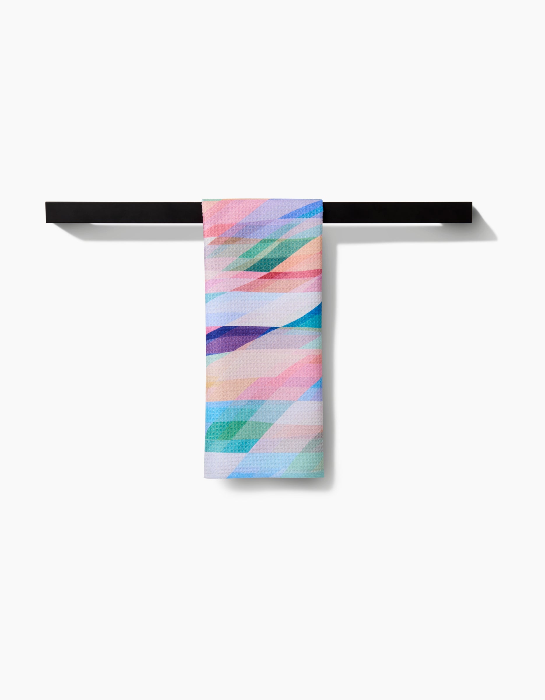 A vibrant Rachel Parker Eventide tea towel with diagonal pink, blue, purple, and teal stripes hangs on a black towel bar against a white background. Crafted from soft, absorbent Geoweave™ fabric.