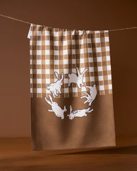The Fall Bunnies tea towel by Geometry features six white rabbits in a circle on a brown and white checkered background, with a solid brown lower section. Made from quick-drying recycled materials, its both stylish and sustainable.