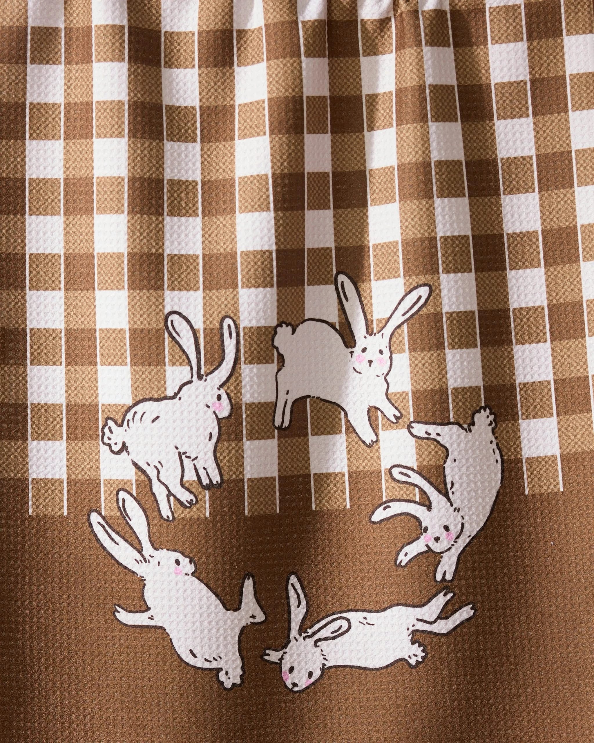 The Geometry Fall Bunnies tea towel features quick-drying recycled fabric in a brown and white checkered pattern, with five white cartoon rabbits with pink cheeks playfully circling at the bottom.