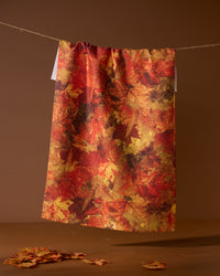 The Geometry Falling Foliage tea towel, featuring an autumn leaf pattern in red, orange, and yellow, hangs on a string against a brown background with scattered leaves below.