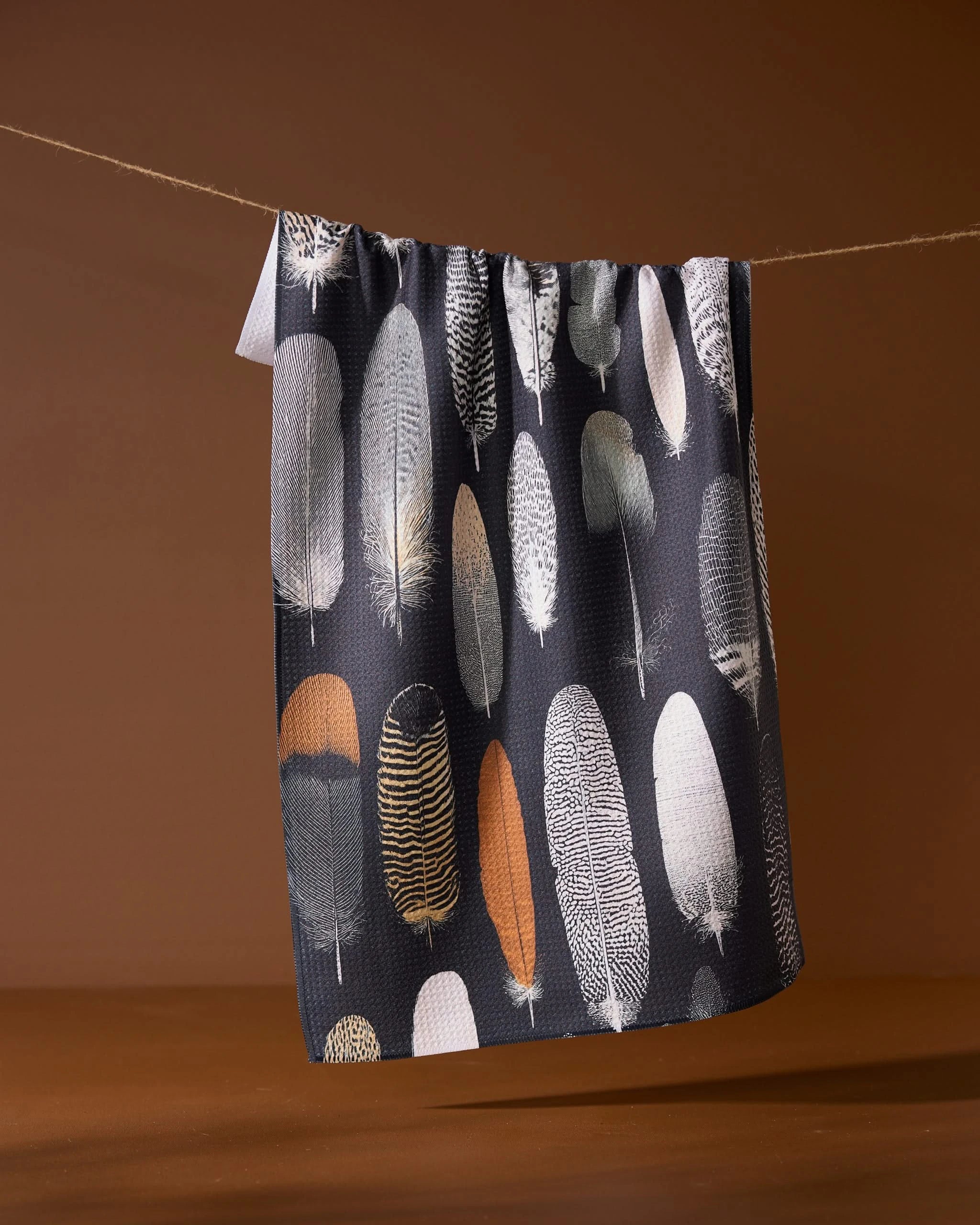 The Geometry Featherfall towel, an eco-friendly, dark-toned piece featuring neutral feather patterns, hangs on a line set against a brown background.