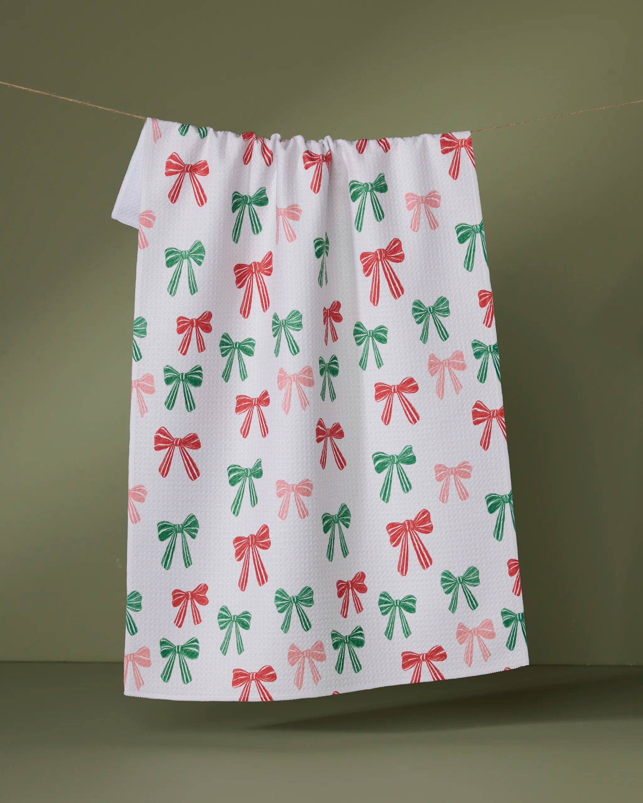 The Geometry Festoon recycled kitchen towel, featuring a red and green bow pattern, hangs on a string against an olive green background.