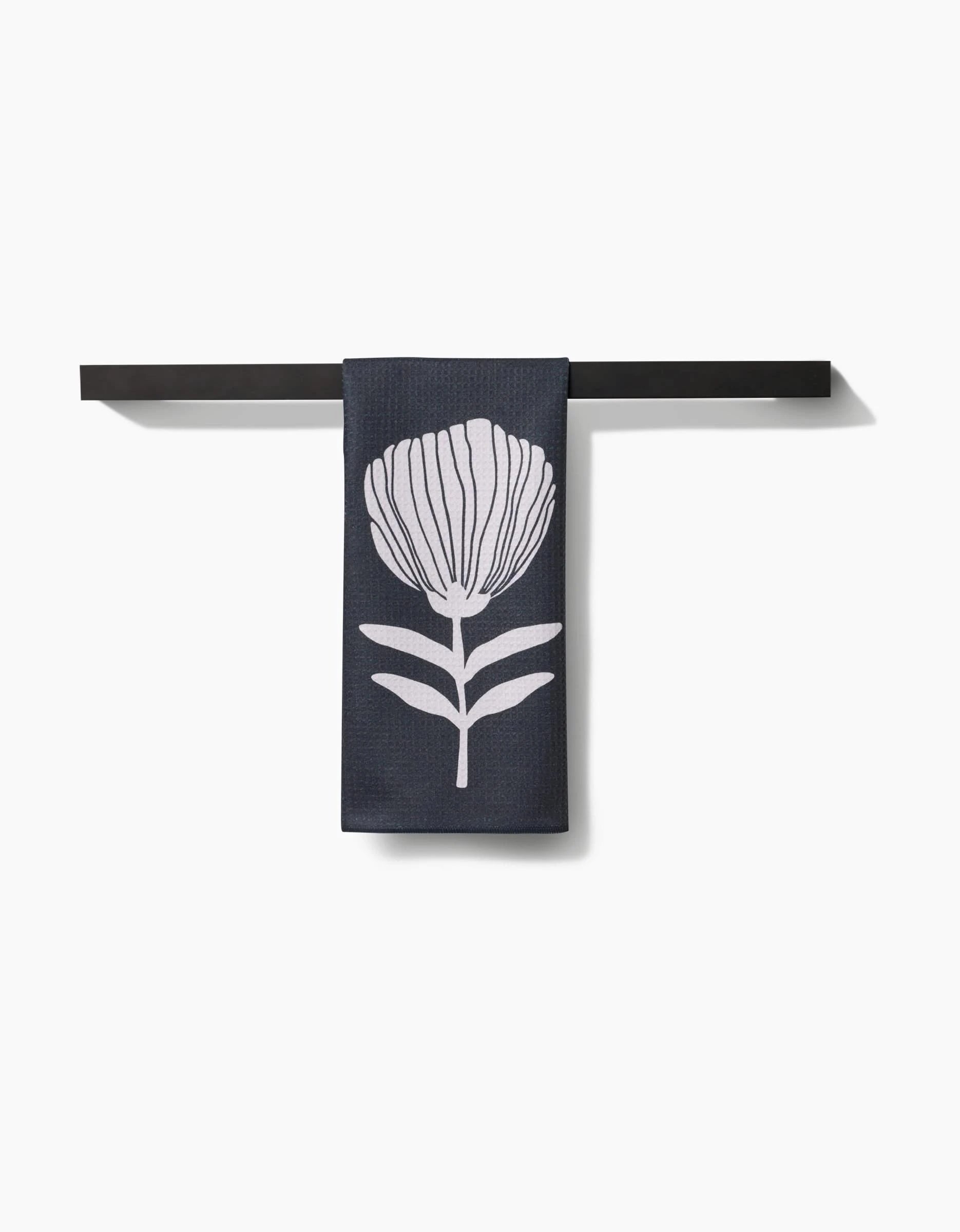 The Anna Christian Fika Tea Towel, featuring a white stylized flower on dark Geoweave™, hangs neatly on a simple black bar against a plain white background.