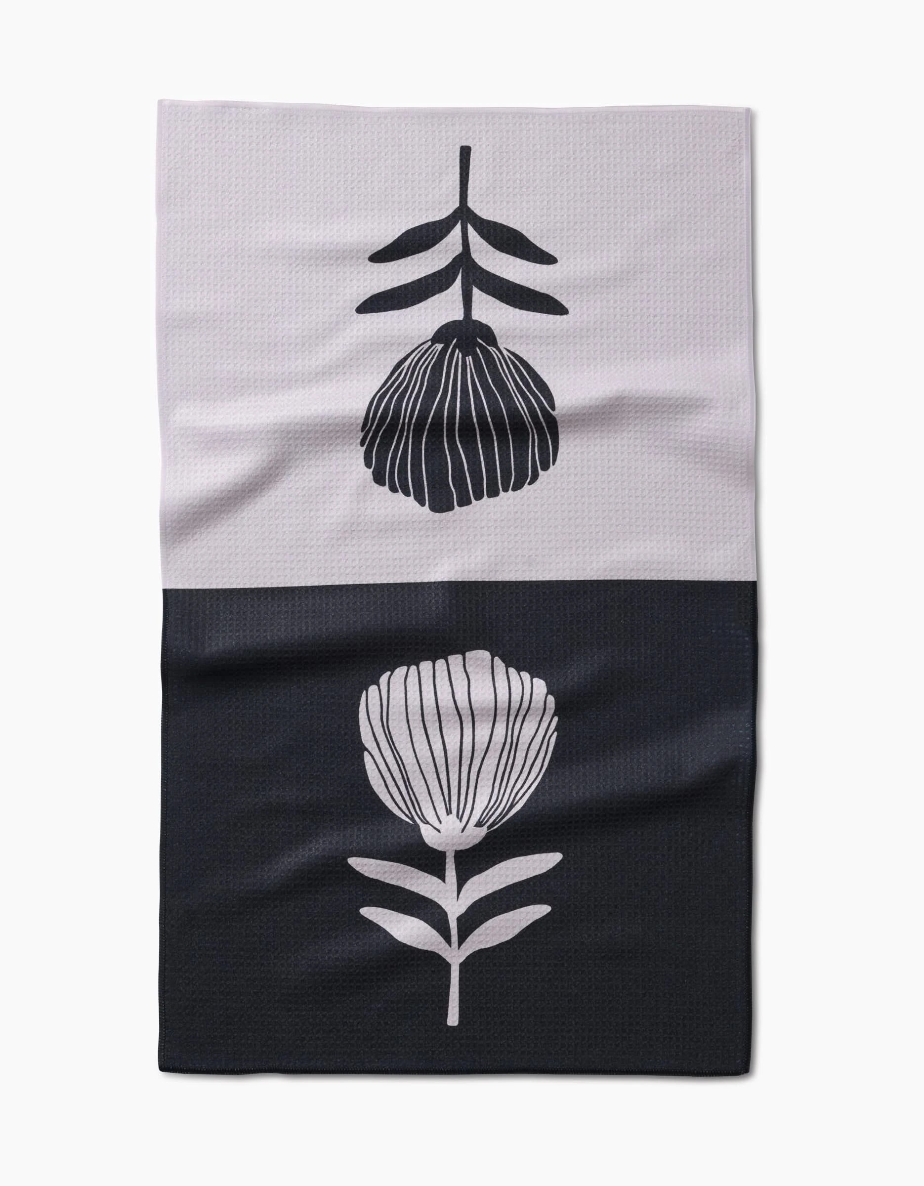 The Fika Tea Towel by Anna Christian features a simple floral design made from recycled materials, with a black flower on light fabric above and a light flower on dark below.