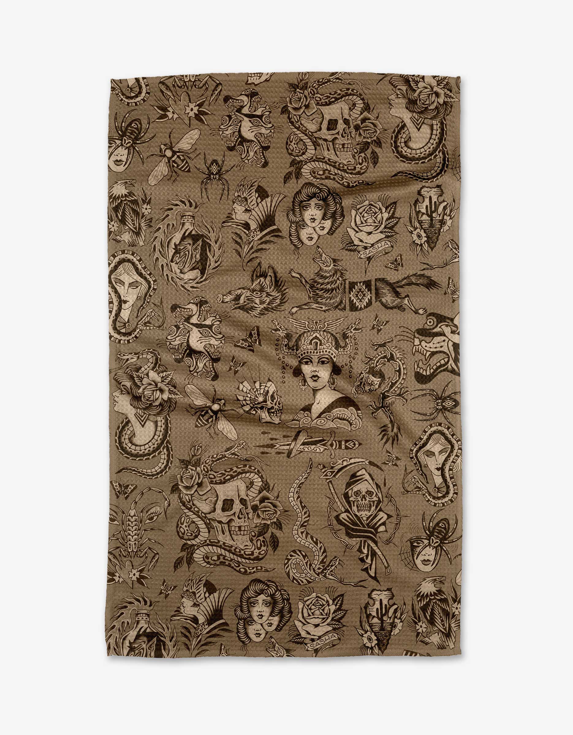 The Flash tea towel by Cole Strem is brown, made from recycled materials, and showcases a dense, repeating pattern of vintage tattoo-style skulls, roses, snakes, women’s faces, and mythical creatures.