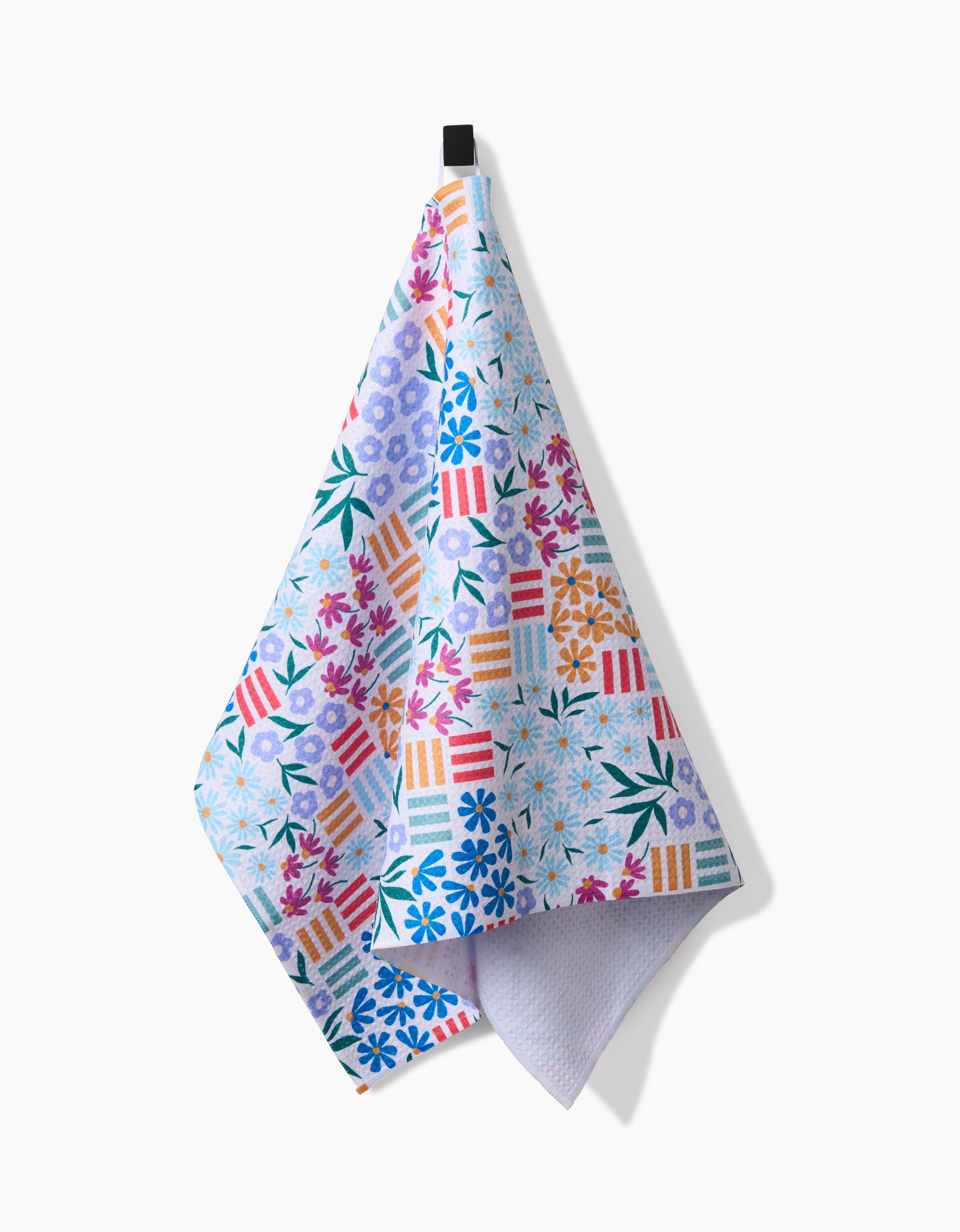 The Floral Garden tea towel by Charlie Rowan Designs, made from recycled materials, features vibrant geometric and floral patterns in blue, red, orange, green, and purple. It hangs on a black hook against a white background.