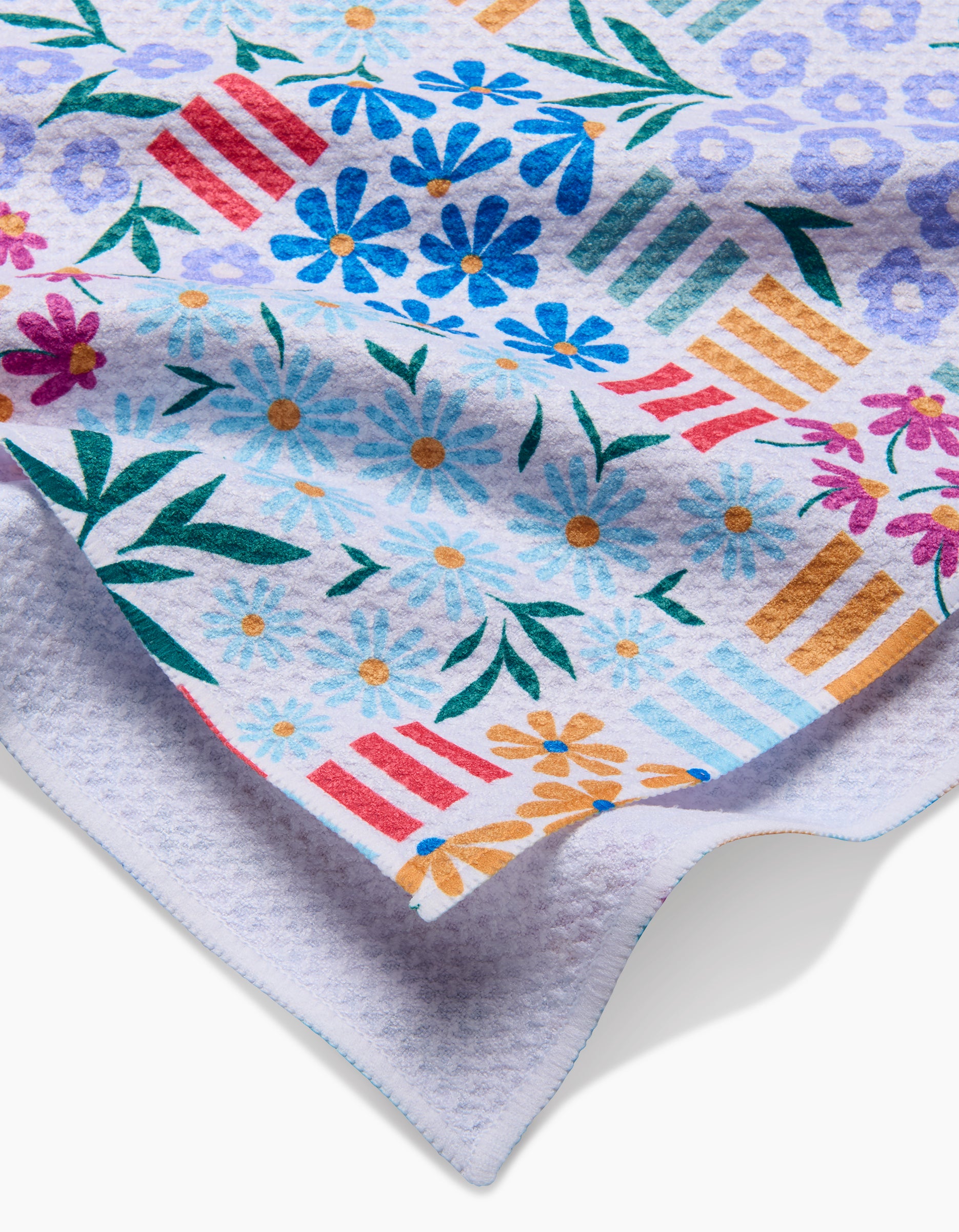 A close-up of the Floral Garden tea towel by Charlie Rowan Designs shows a vibrant floral and striped pattern on white, with a curled edge revealing its textured Geoweave fabric underside.