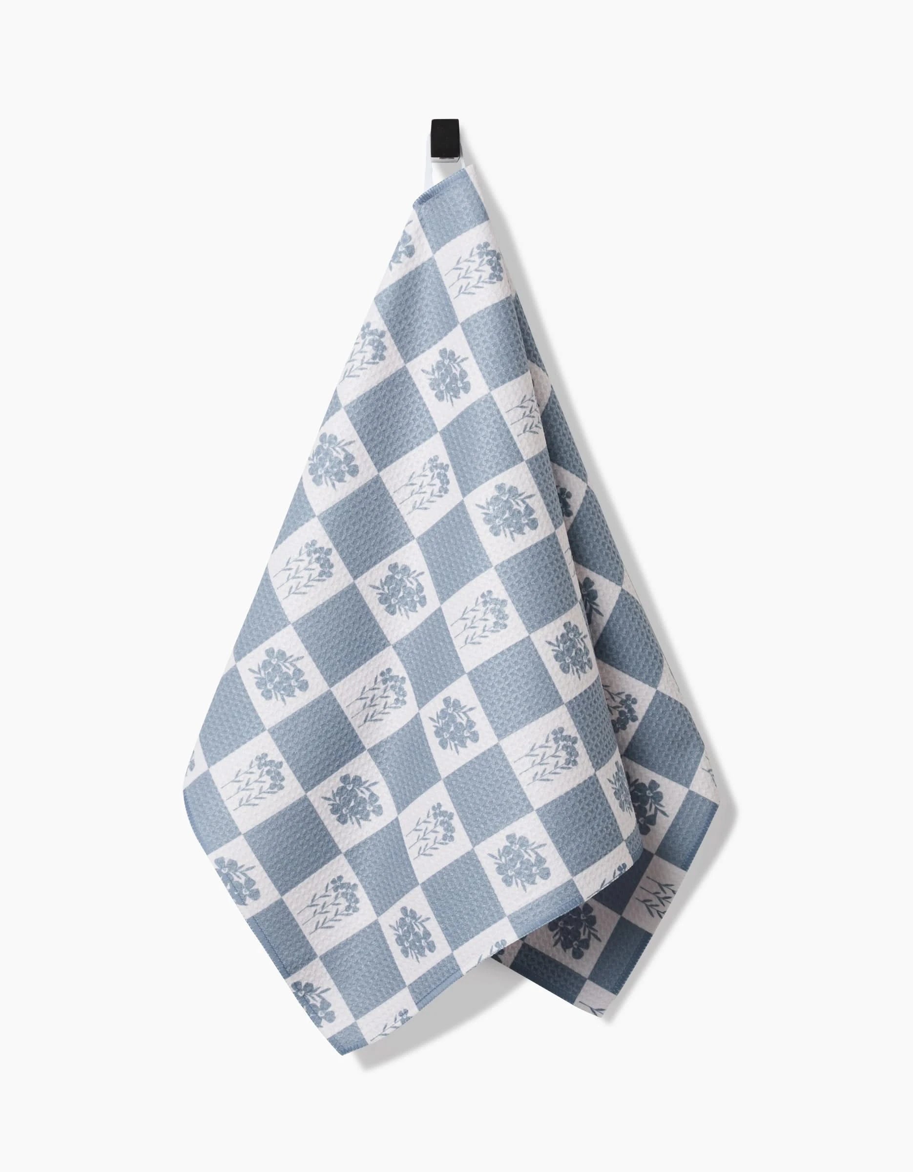 The Floral Patchwork tea towel by Charlie Rowan Designs, featuring a blue and white checkered diamond and floral pattern, crafted from recycled materials, hangs from a black loop against a white background.