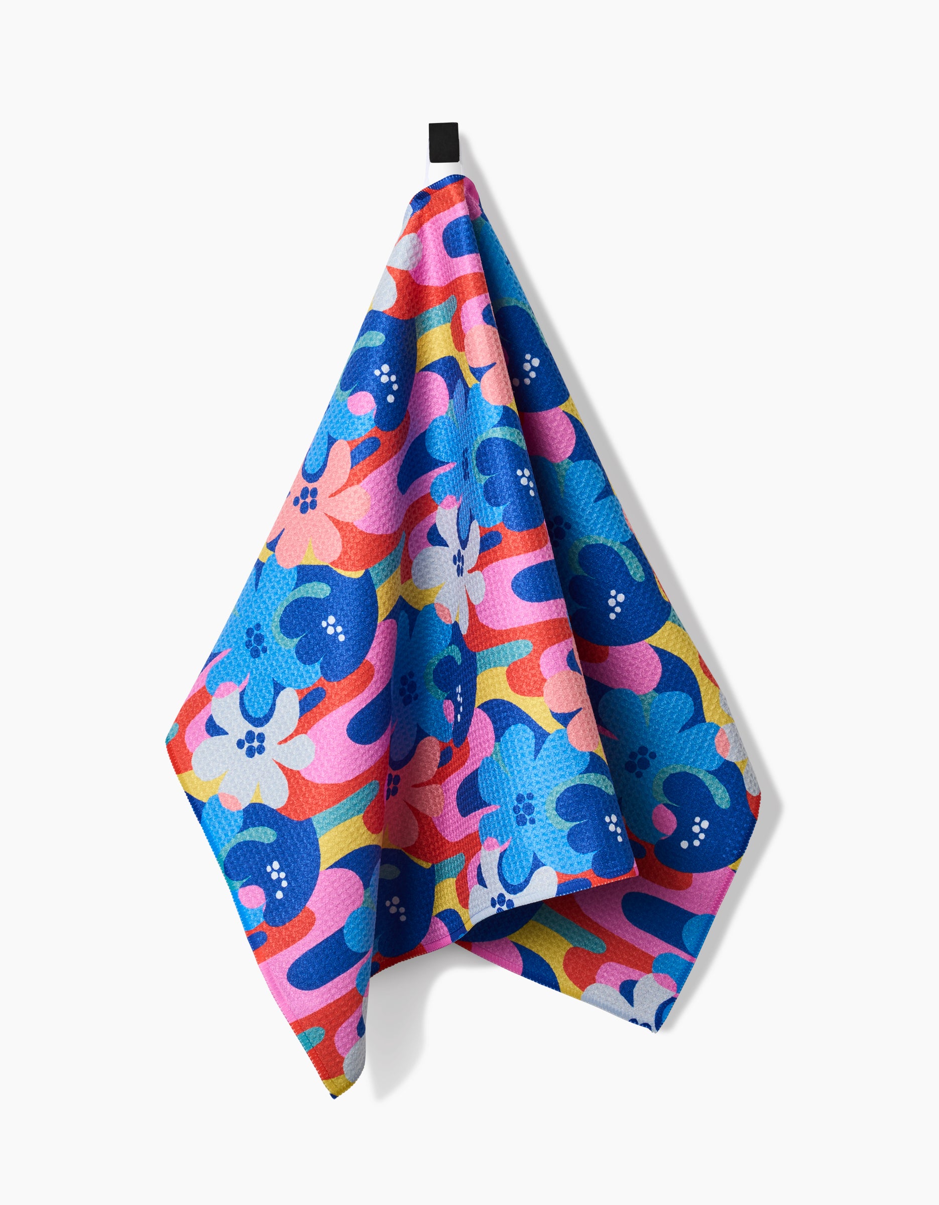 The Flower Powers tea towel by Masha Volnova features a bold floral pattern in blue, pink, orange, yellow, and white. Crafted with Geoweave™ fabric for exceptional absorbency and durability; hangs from a black loop on a white background.