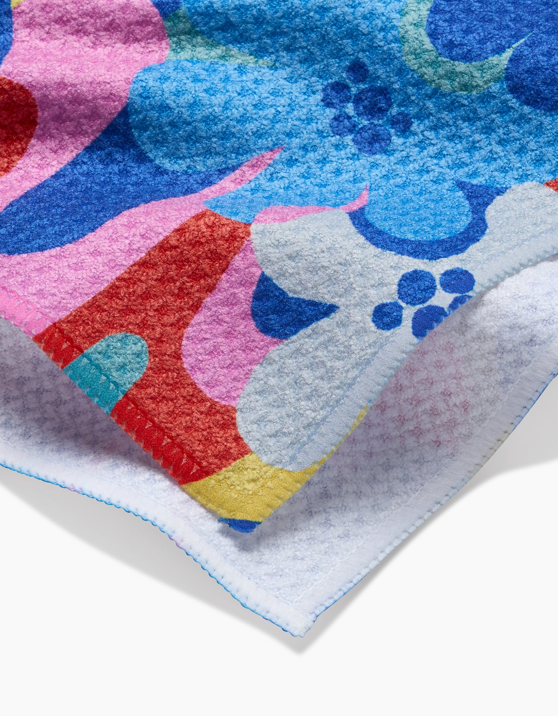 A close-up of the Flower Powers tea towel by Masha Volnova shows its textured Geoweave™ fabric with large, overlapping blue, red, pink, and white flowers and a visible edge finished with blue stitching.