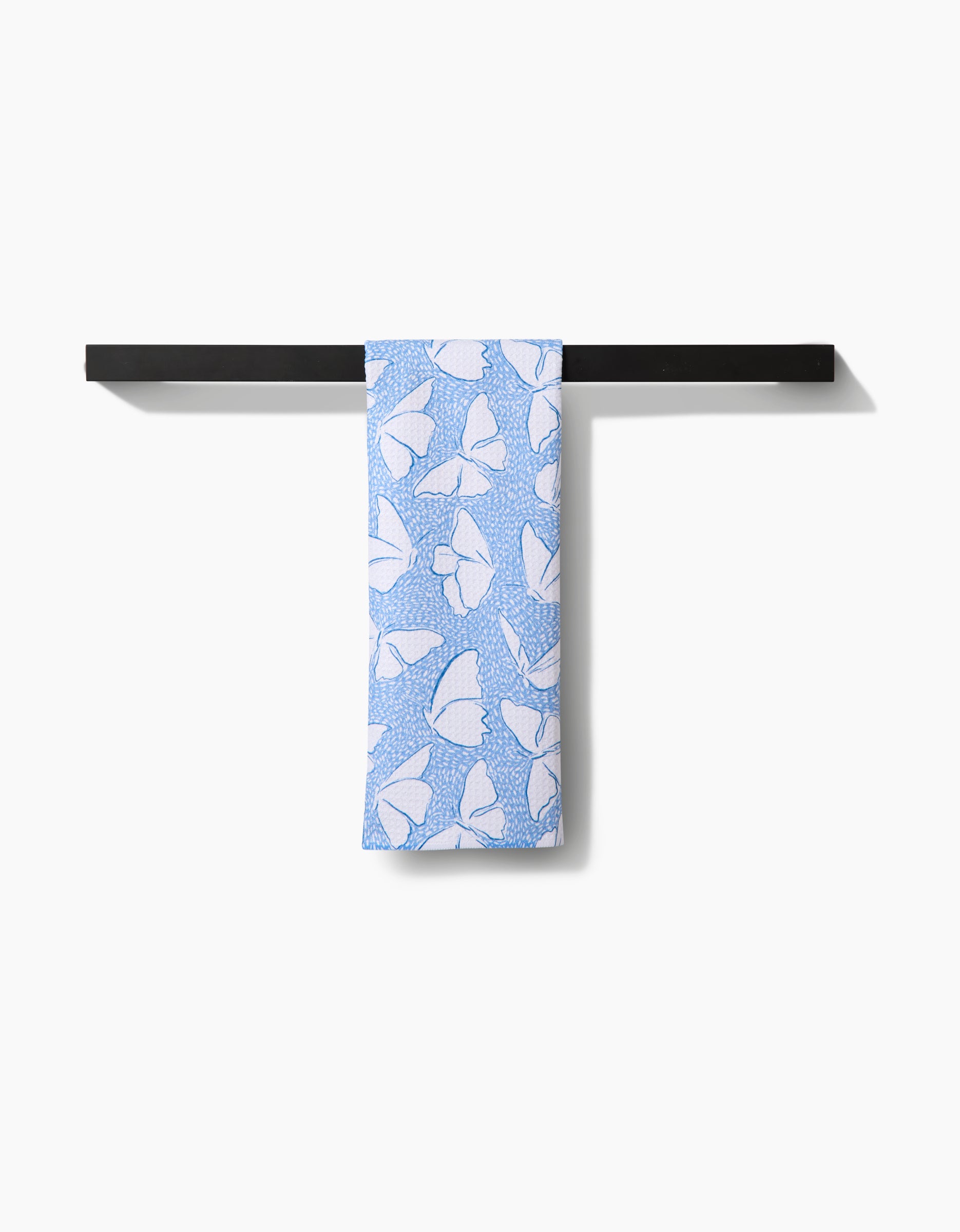 The Hufton Studio Fly Away Butterfly tea towel, featuring a white butterfly pattern on blue Geoweave fabric made from recycled materials, hangs neatly from a black towel bar against a white wall—an eco-friendly choice.