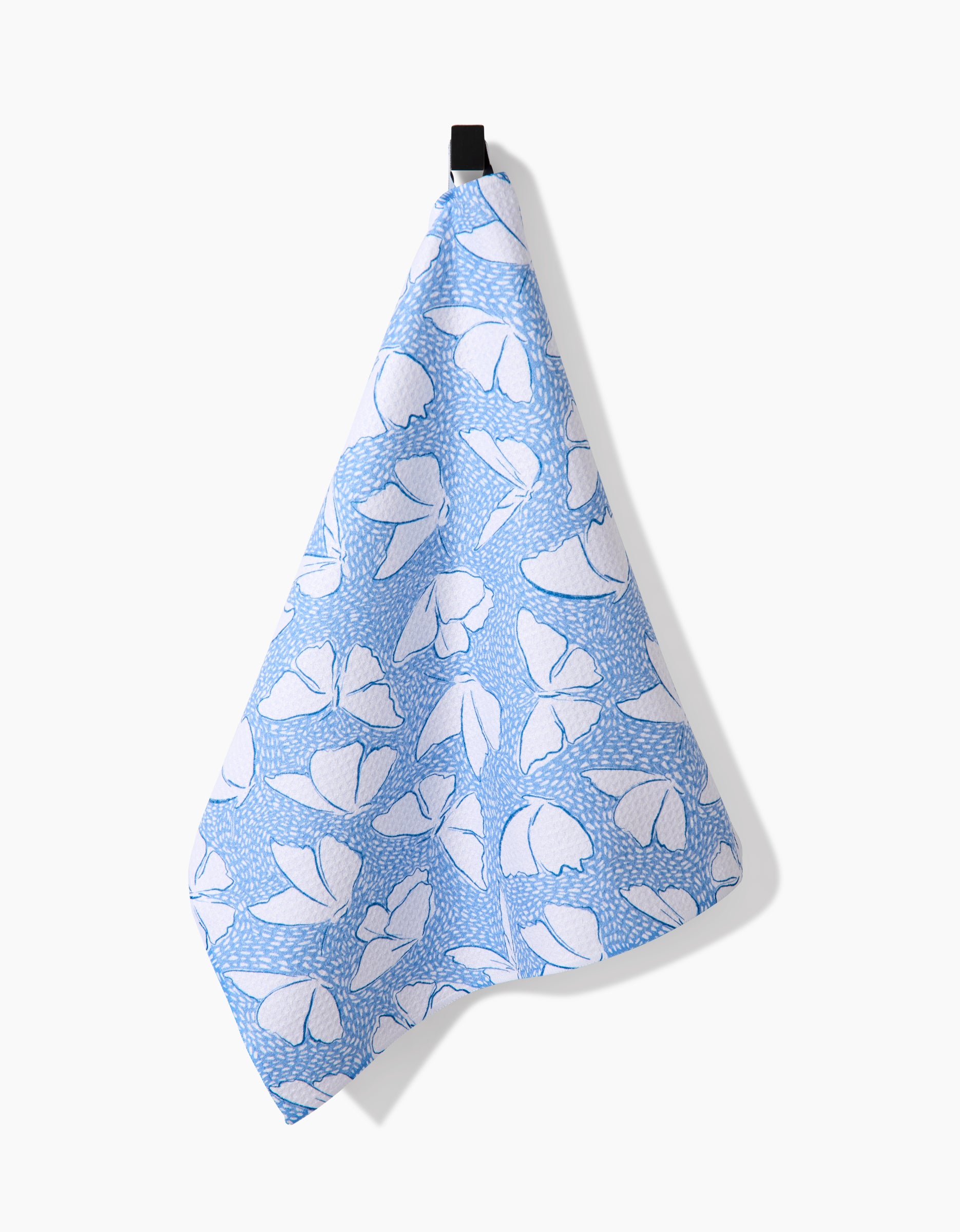 A blue and white Fly Away Butterfly tea towel by Hufton Studio, featuring an abstract butterfly design and made from recycled materials, hangs from a black loop against a plain white background.