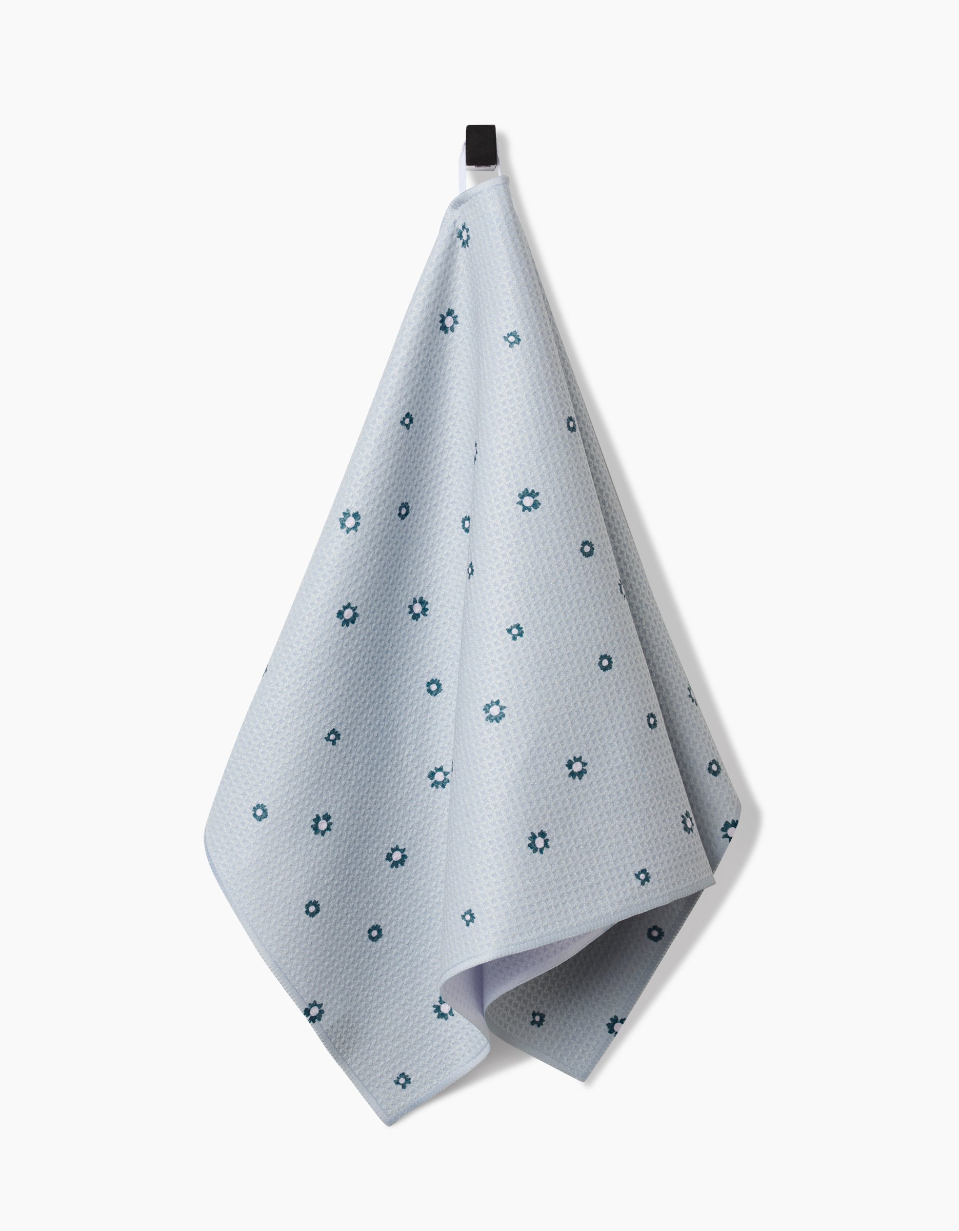 The Forest Daisy tea towel by Coit Creative hangs from a black hook against a white background, featuring a subtle pattern of small dark blue florals on light blue Geoweave™ fabric.