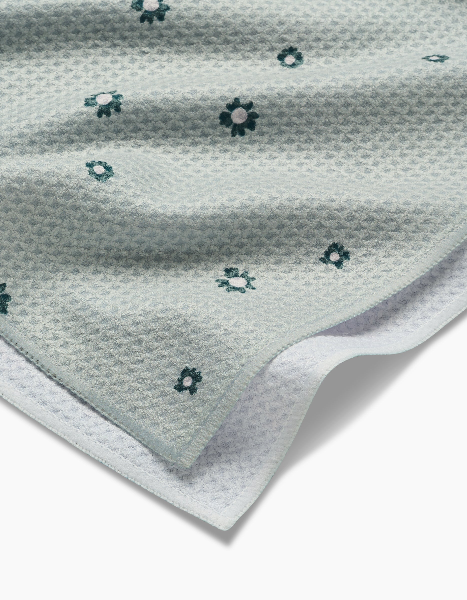 The Forest Daisy tea towel by Coit Creative features light blue Geoweave™ fabric with a waffle weave pattern and small embroidered dark green and white flowers, shown partially folded on a white background.