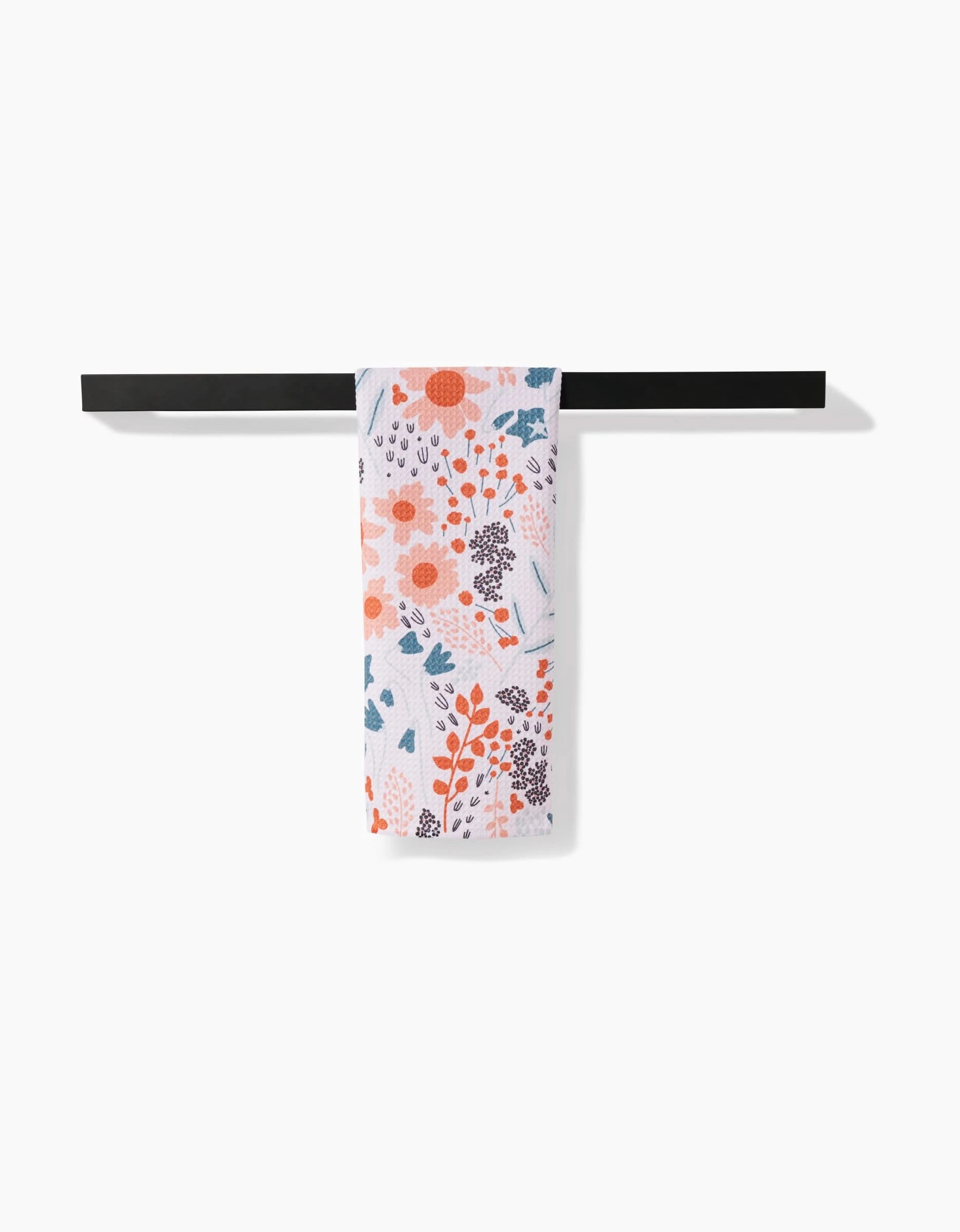 The Coit Creative Forest Foliage tea towel, made from Geoweave fabric and featuring an orange, red, and blue floral pattern, hangs neatly on a black towel bar against a white background.