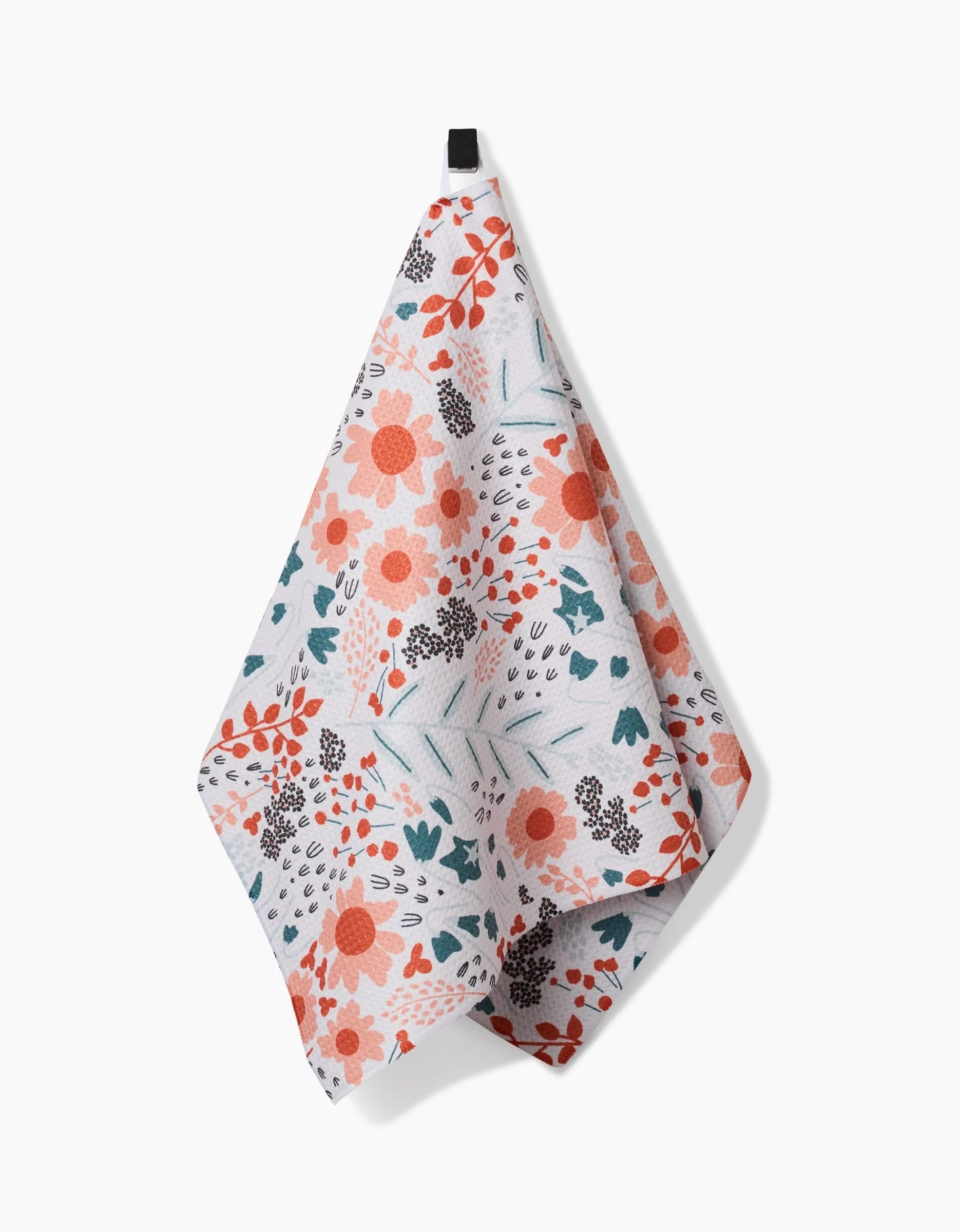The Forest Foliage tea towel by Coit Creative hangs from a black loop and features a floral print in orange, pink, blue, and green on white Geoweave fabric.