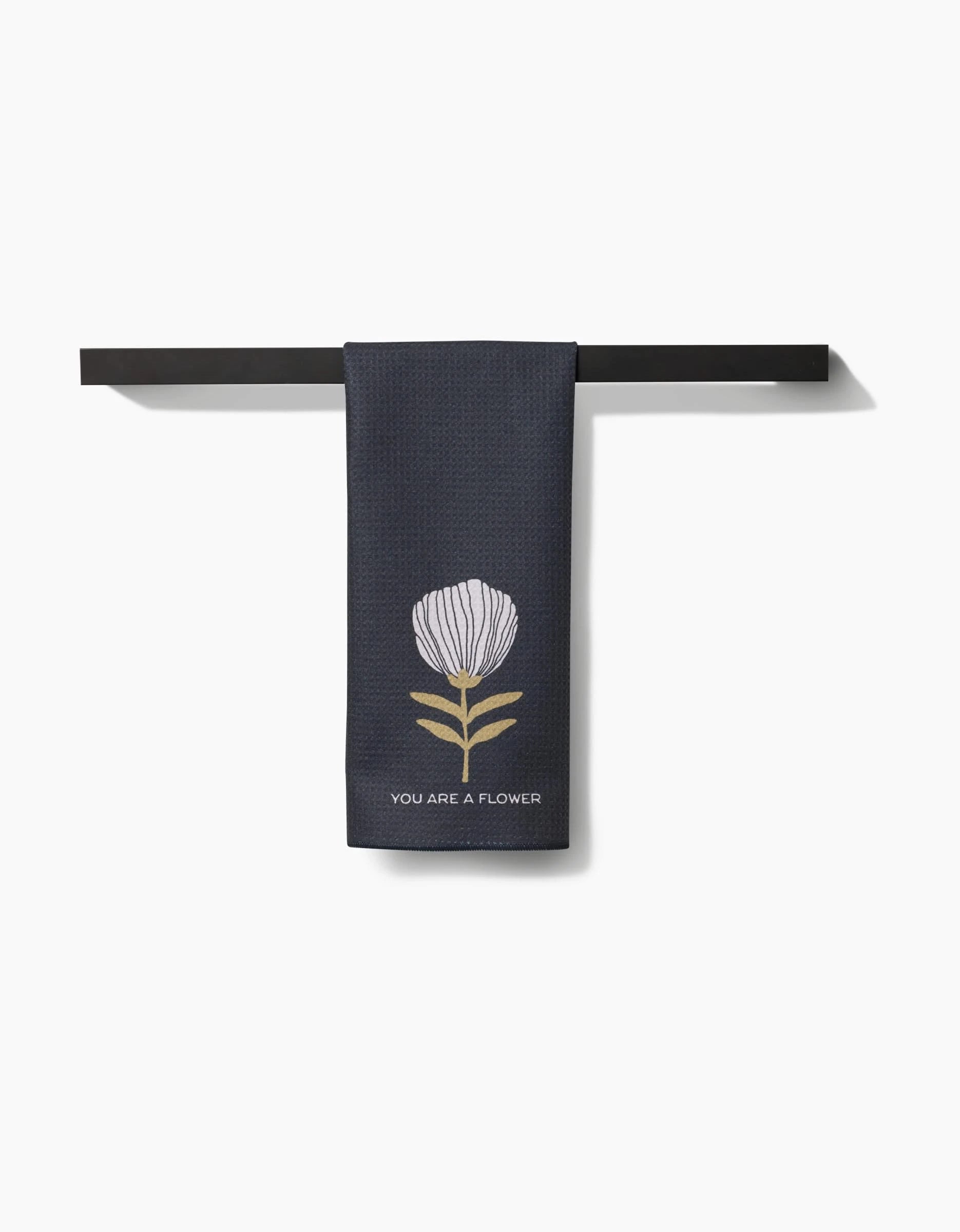 The Freja tea towel by Anna Christian is dark blue Geoweave fabric, hangs on a black rod, and displays a white flower with green leaves plus YOU ARE A FLOWER below the image.