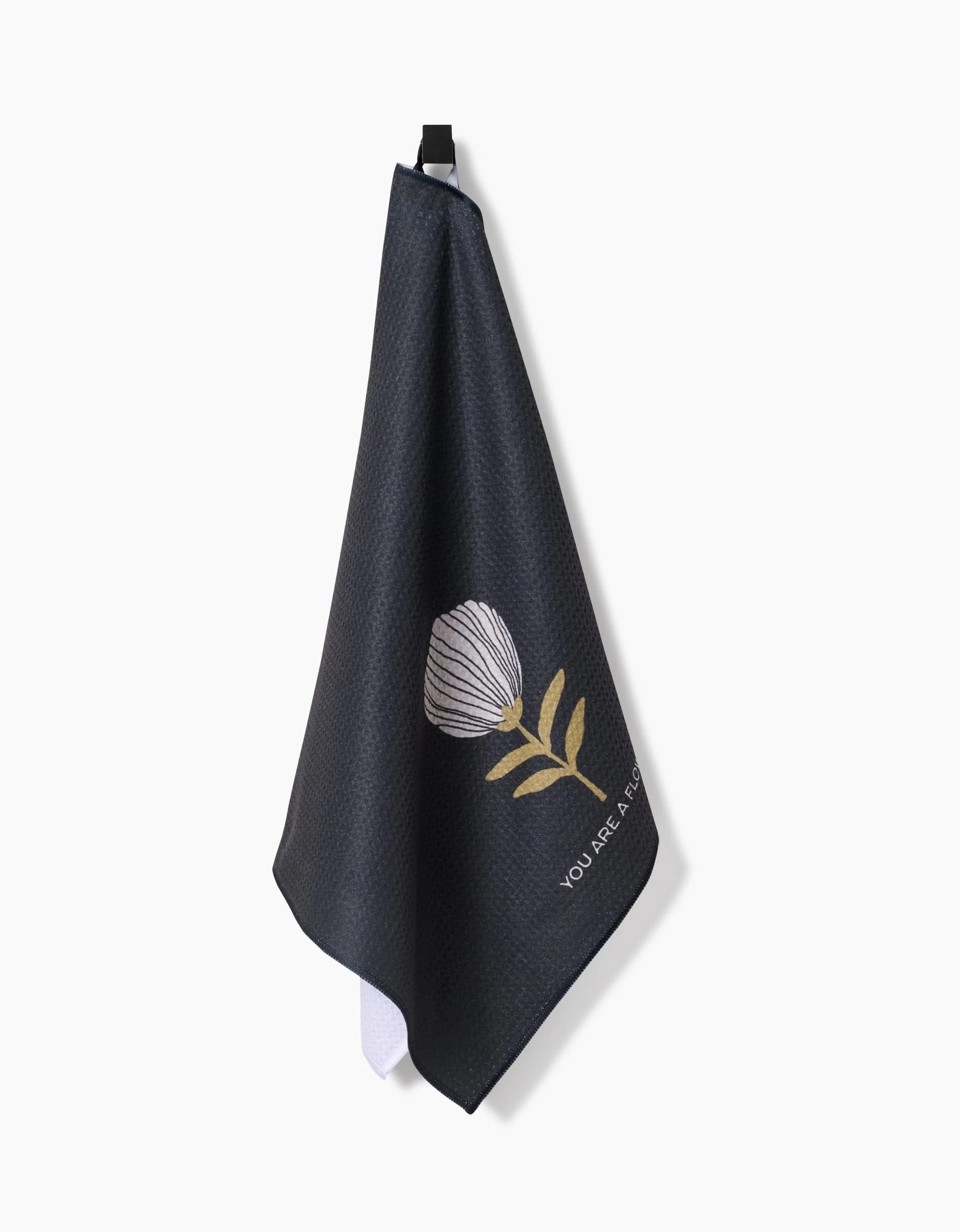 The Anna Christian Freja tea towel features dark Geoweave fabric, a hanging loop, a simple white and yellow floral design, and “YOU ARE ENOUGH” printed on one corner.