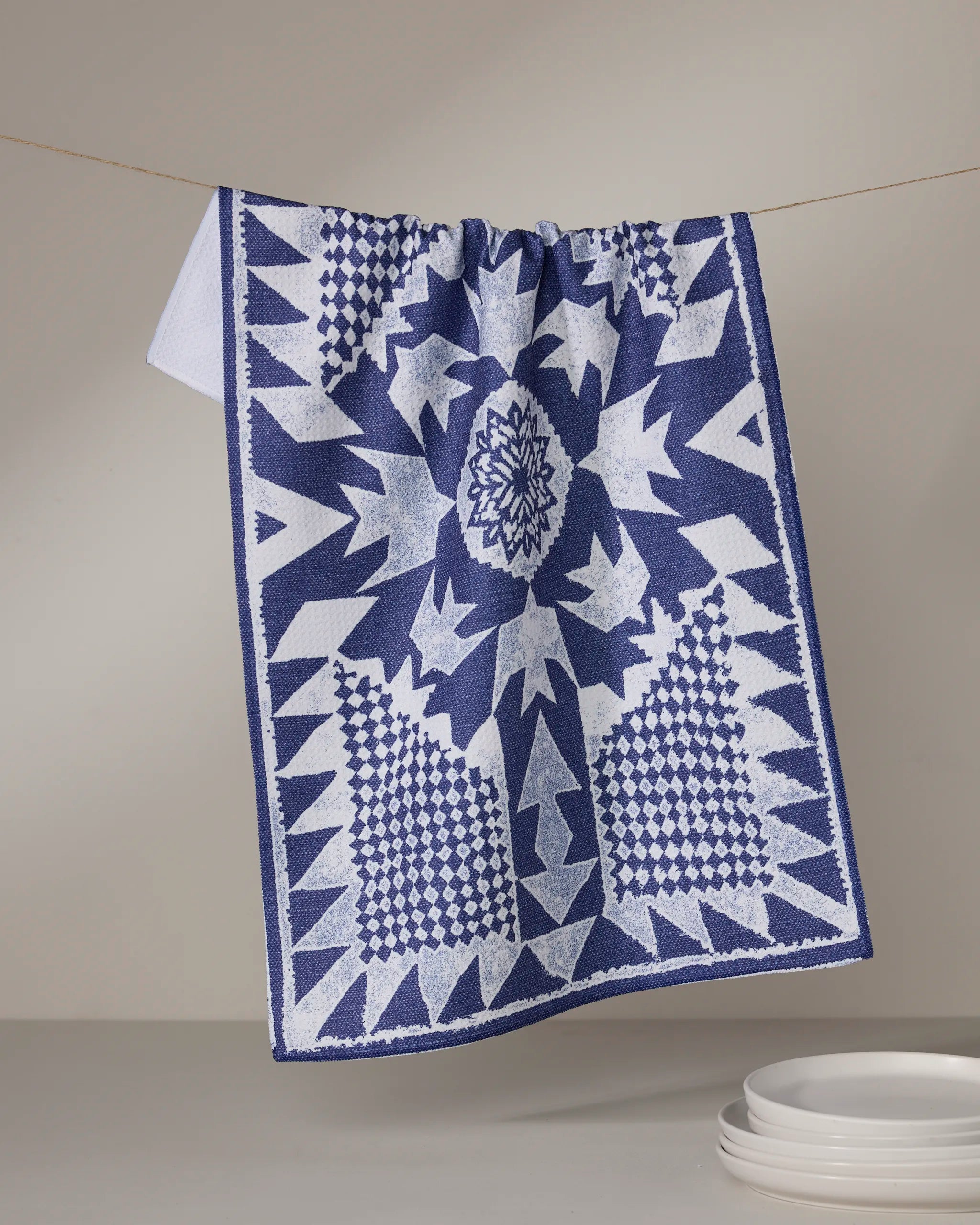 The Geometry Froststar kitchen towel, featuring blue and white geometric star patterns, hangs above a stack of white plates against a neutral background.